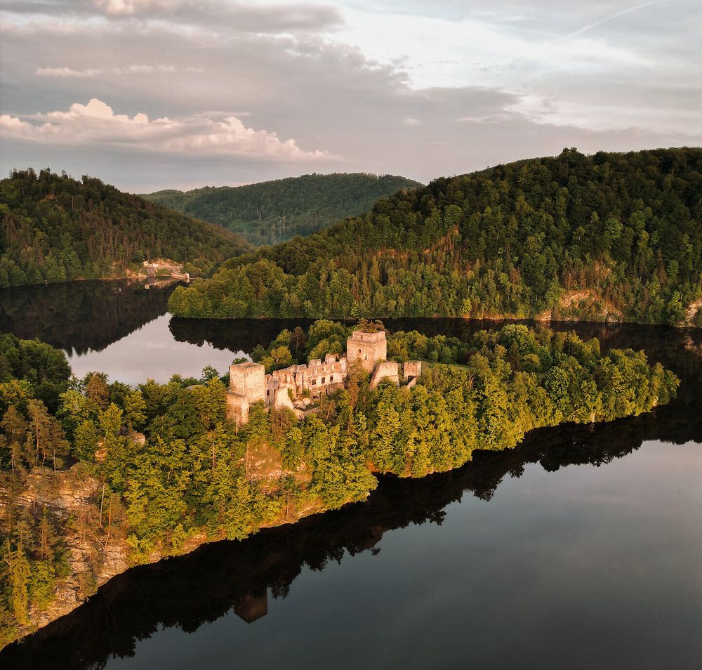 Eine Luftaufnahme der Burgruine Dobra im Waldviertel, umgeben von dicht bewaldeten Hügeln und einem See bei Sonnenuntergang.