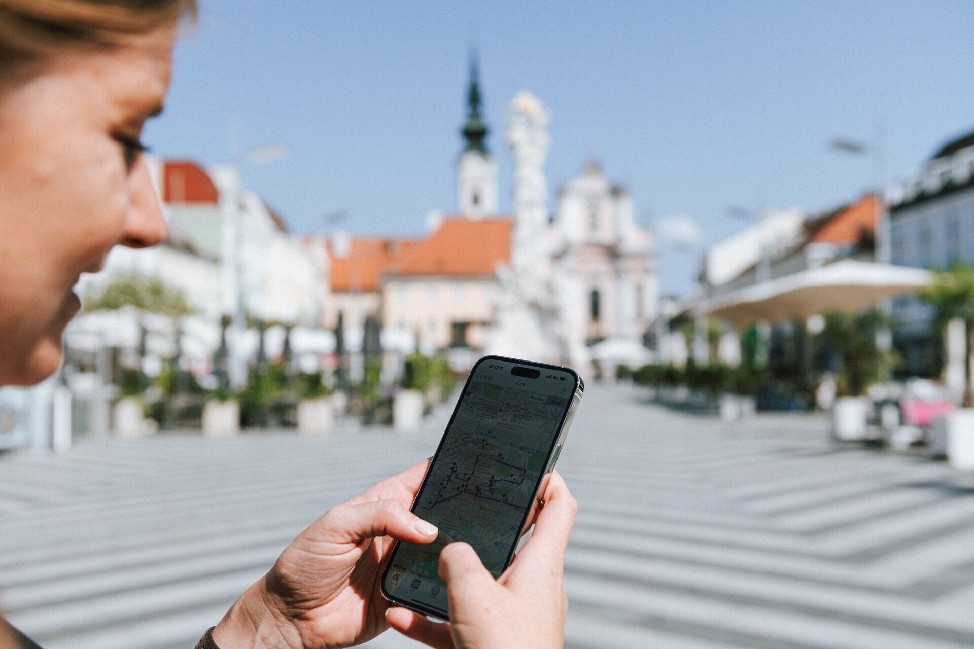 An einem sonnigen Tag in der Stadt wird der Rathausplatz lebendig. Eine Person betrachtet auf ihrem Handy die Umgebung und plant ihren nächsten Schritt, während die beeindruckende Architektur im Hintergrund strahlt. Die Atmosphäre ist einladend und voller Möglichkeiten für Erkundungen.