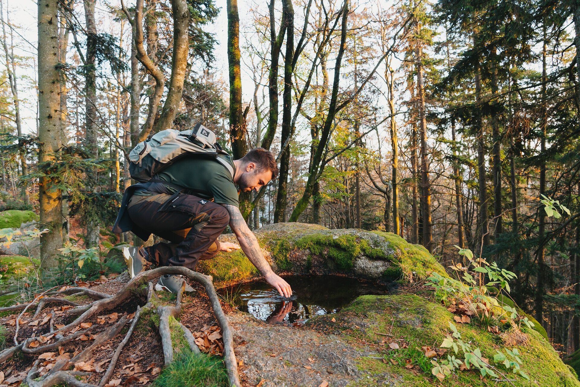 In der malerischen Ysperklamm, umgeben von majestätischen Bäumen und sanften Hügeln, kniet ein Wanderer am Ufer eines klaren Baches. Die goldenen Herbstblätter reflektieren das sanfte Licht der Nachmittagssonne und schaffen eine friedliche Atmosphäre, die zum Verweilen einlädt.