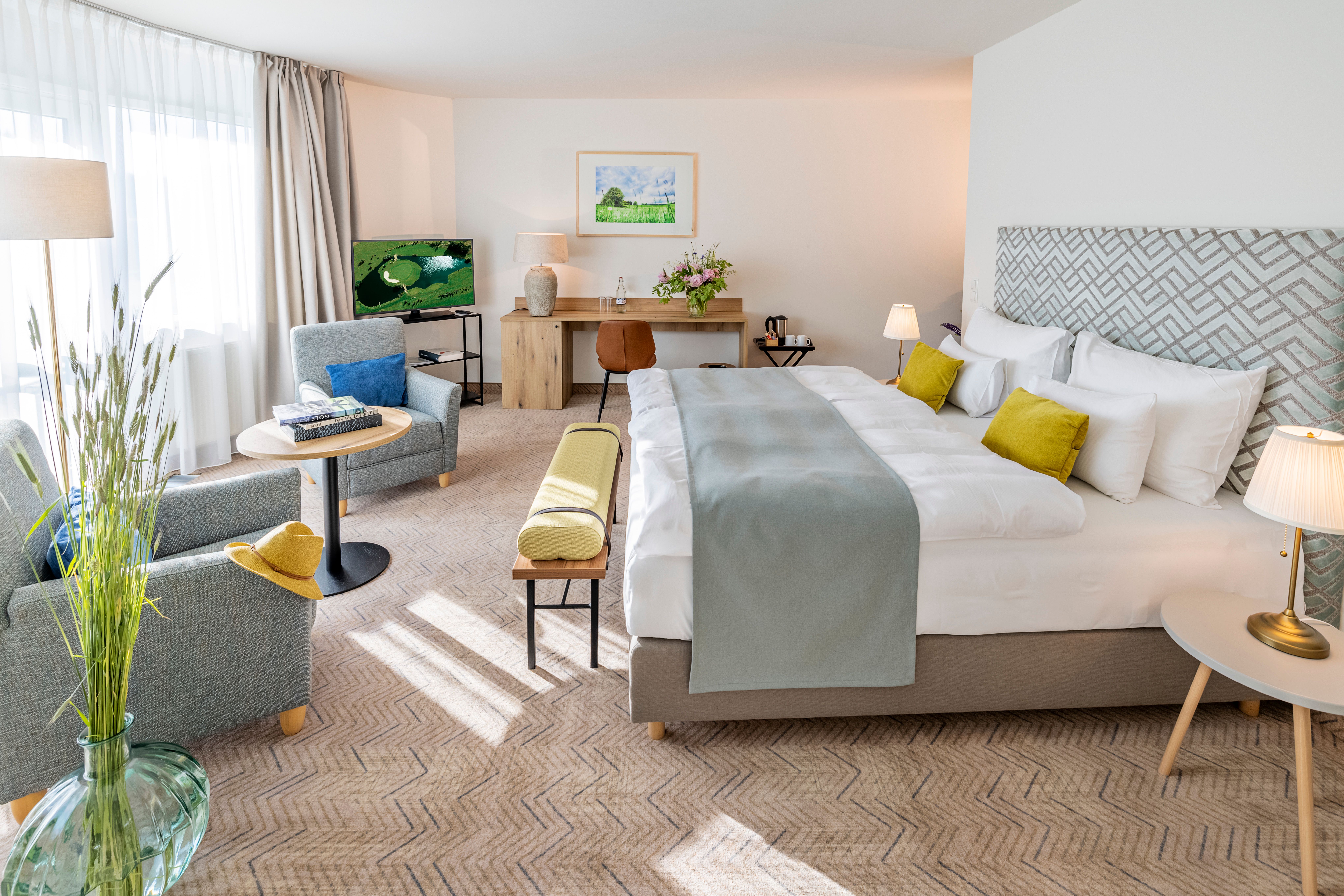 Helles Hotelzimmer im Golfresort Haugschlag mit Doppelbett, Sessel, Schreibtisch, TV und großen Fenstern.