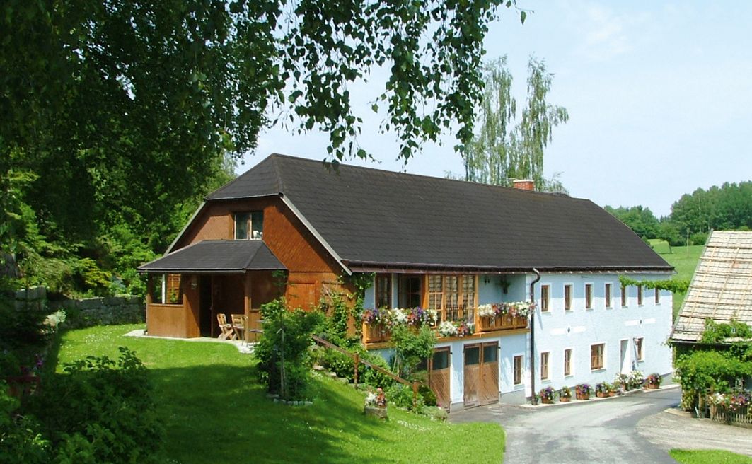 Ein traditionelles Bauernhaus mit Blumen auf dem Balkon, umgeben von grüner Landschaft und Bäumen.