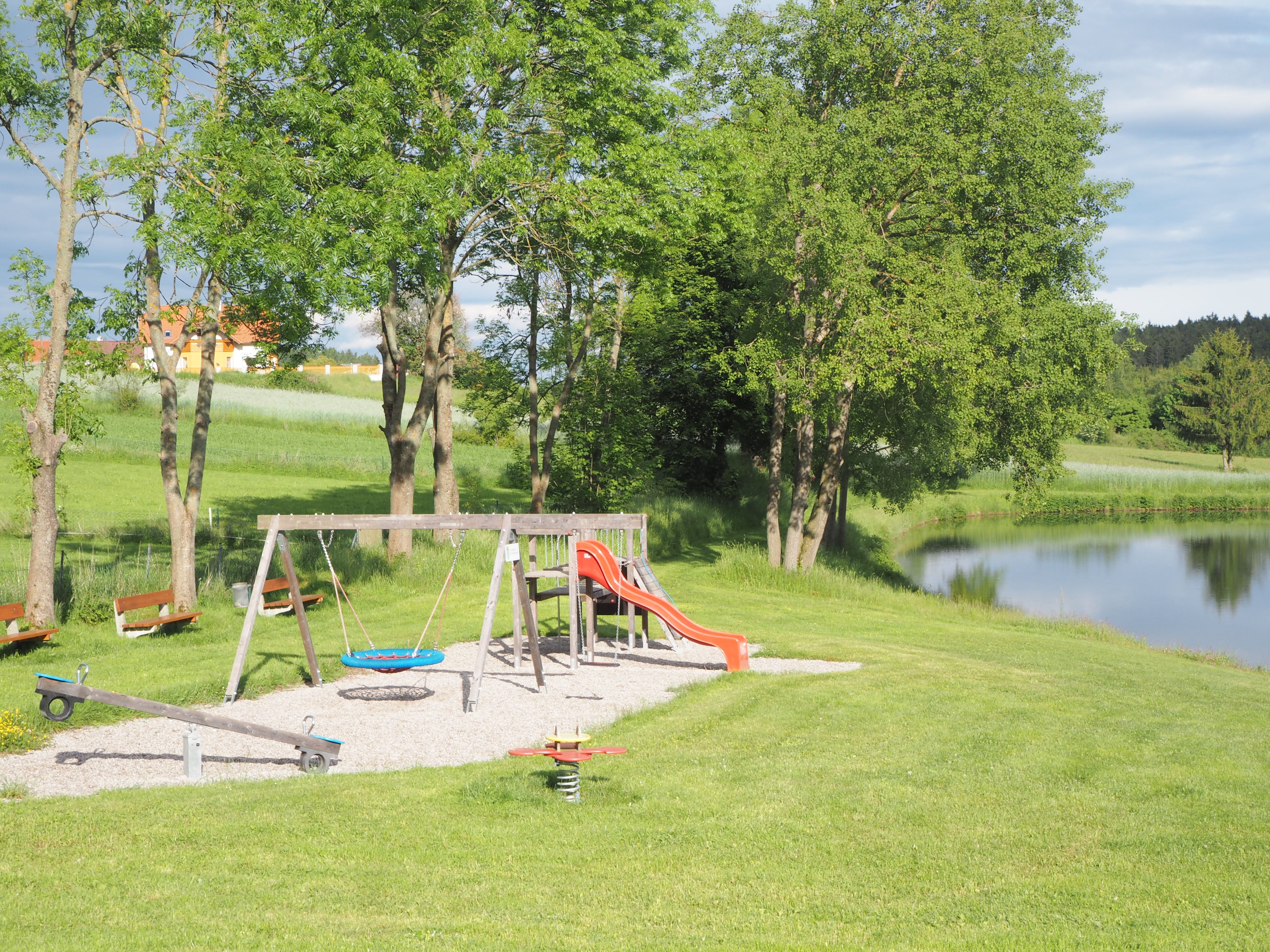 Spielplatz mit Schaukel, Rutsche und Wippe neben einem Teich in einer grünen Landschaft.