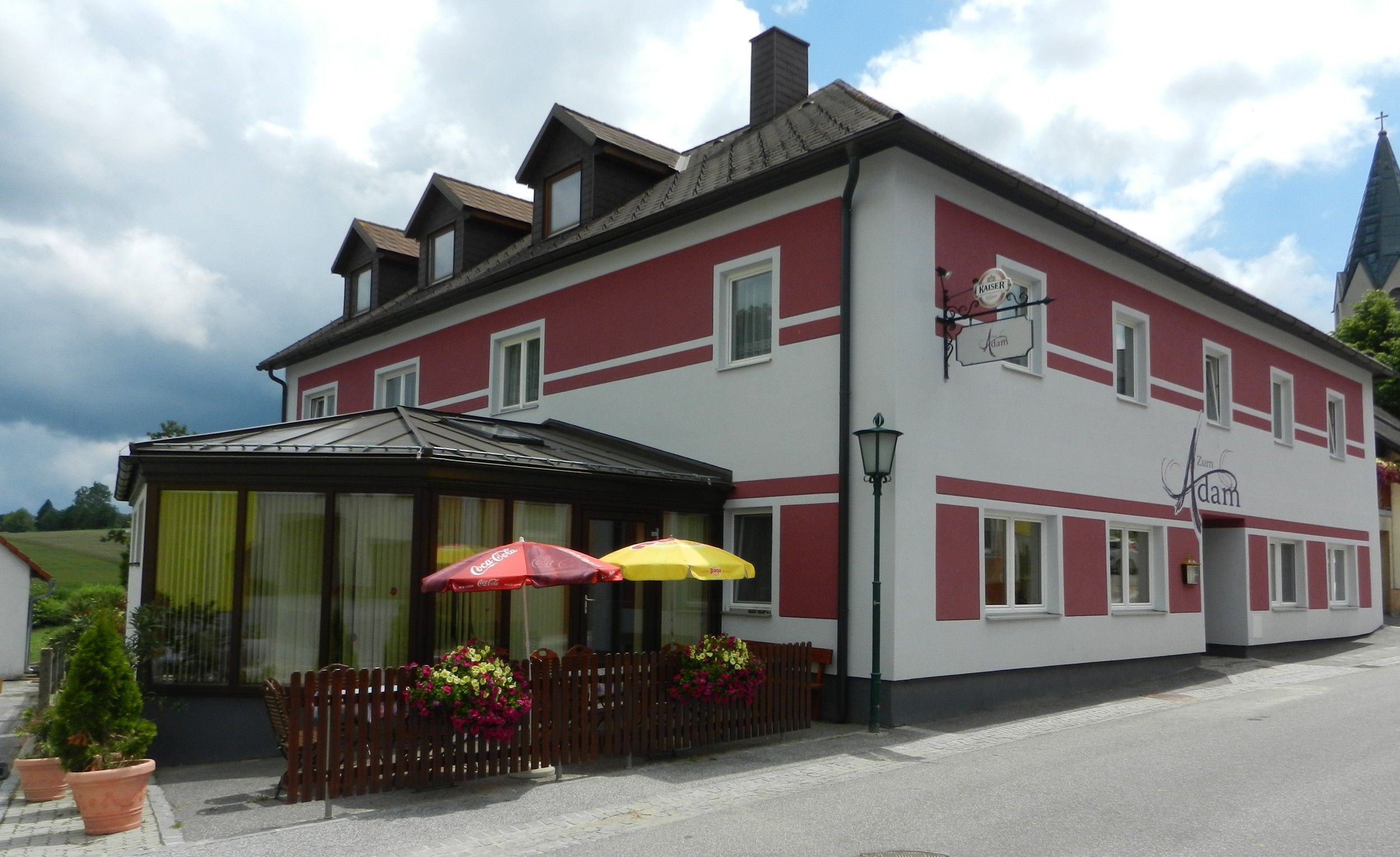 Ein Gasthaus mit roter und weißer Fassade, Terrasse mit Sonnenschirmen und Blumendekoration.