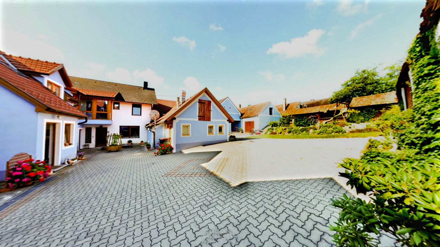 Hof mit mehreren Gebäuden, blauer Himmel, gepflasterter Boden, Blumen und Pflanzen.