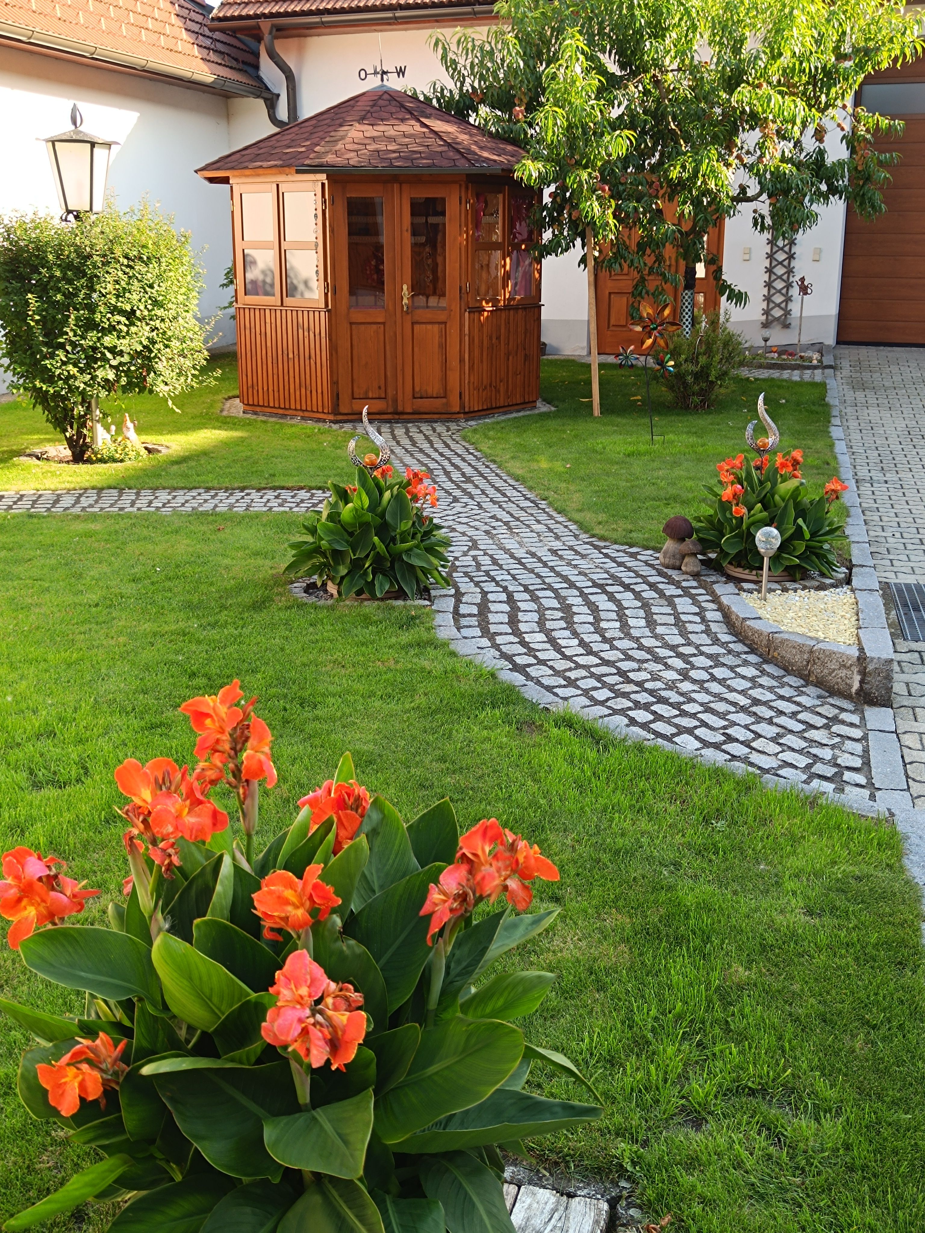 Ein gepflegter Garten mit einem kleinen Holzpavillon, umgeben von grüner Wiese und blühenden orangefarbenen Blumen.