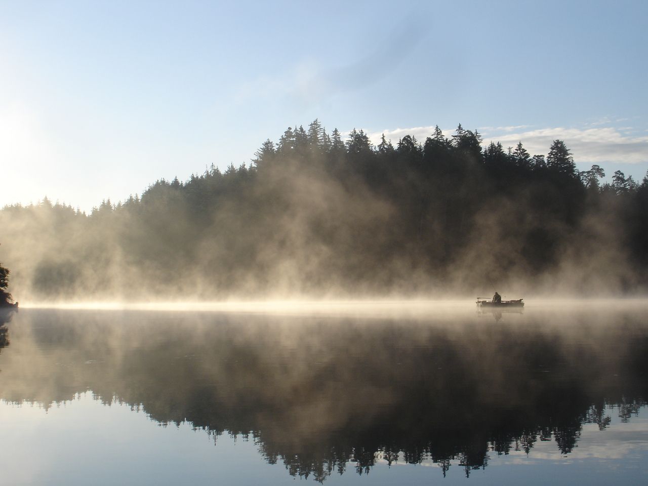 Ein Boot auf einem nebligen See vor einem bewaldeten Ufer bei Sonnenaufgang.