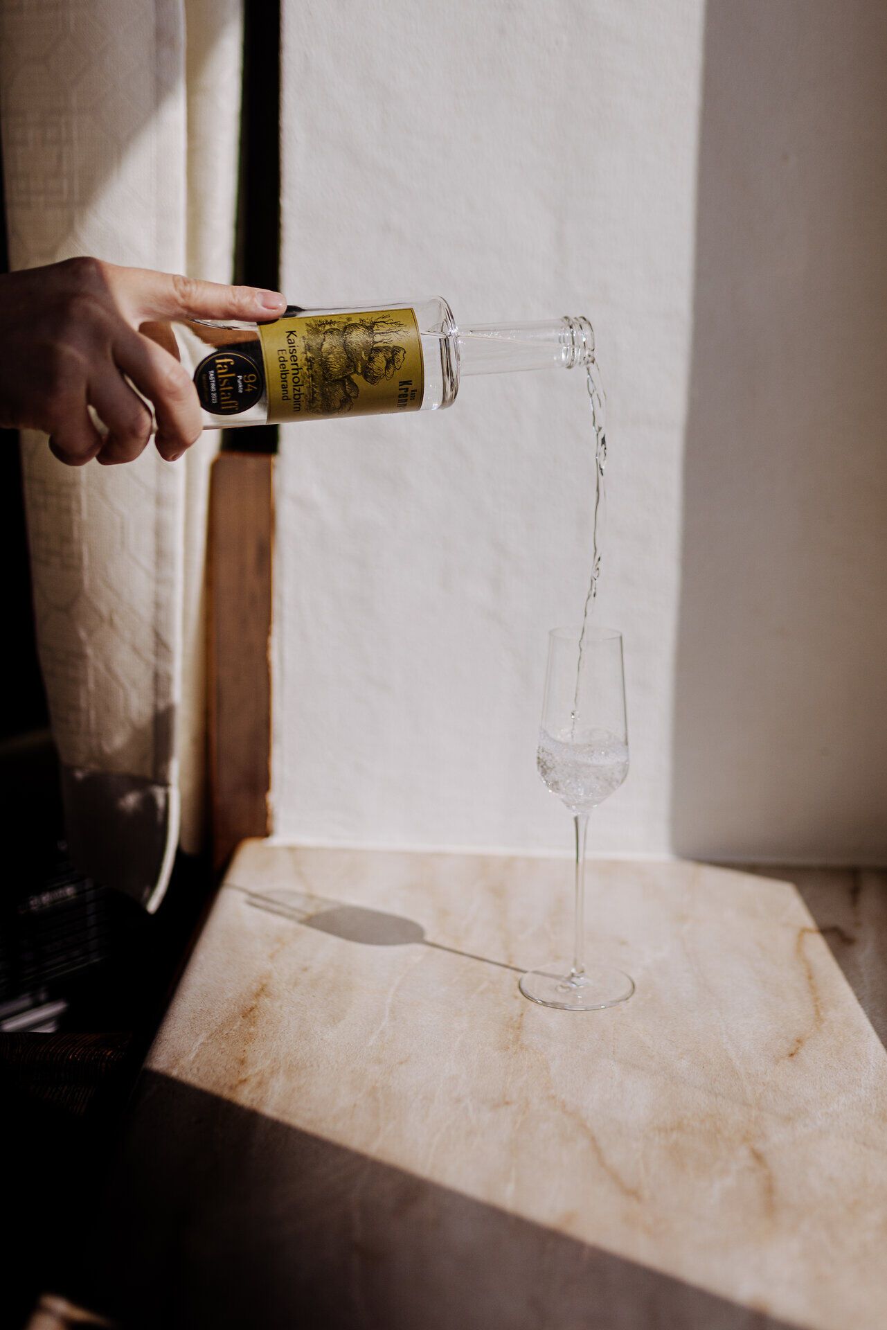 Ein strahlender Sonnenstrahl fällt durch das Fenster und beleuchtet das elegante Glas, während der klare Edelbrand sanft hineinfließt. Die Atmosphäre ist geprägt von der Vorfreude auf genussvolle Momente, die hier in der Wirtshausbrennerei Krenn erlebt werden können.