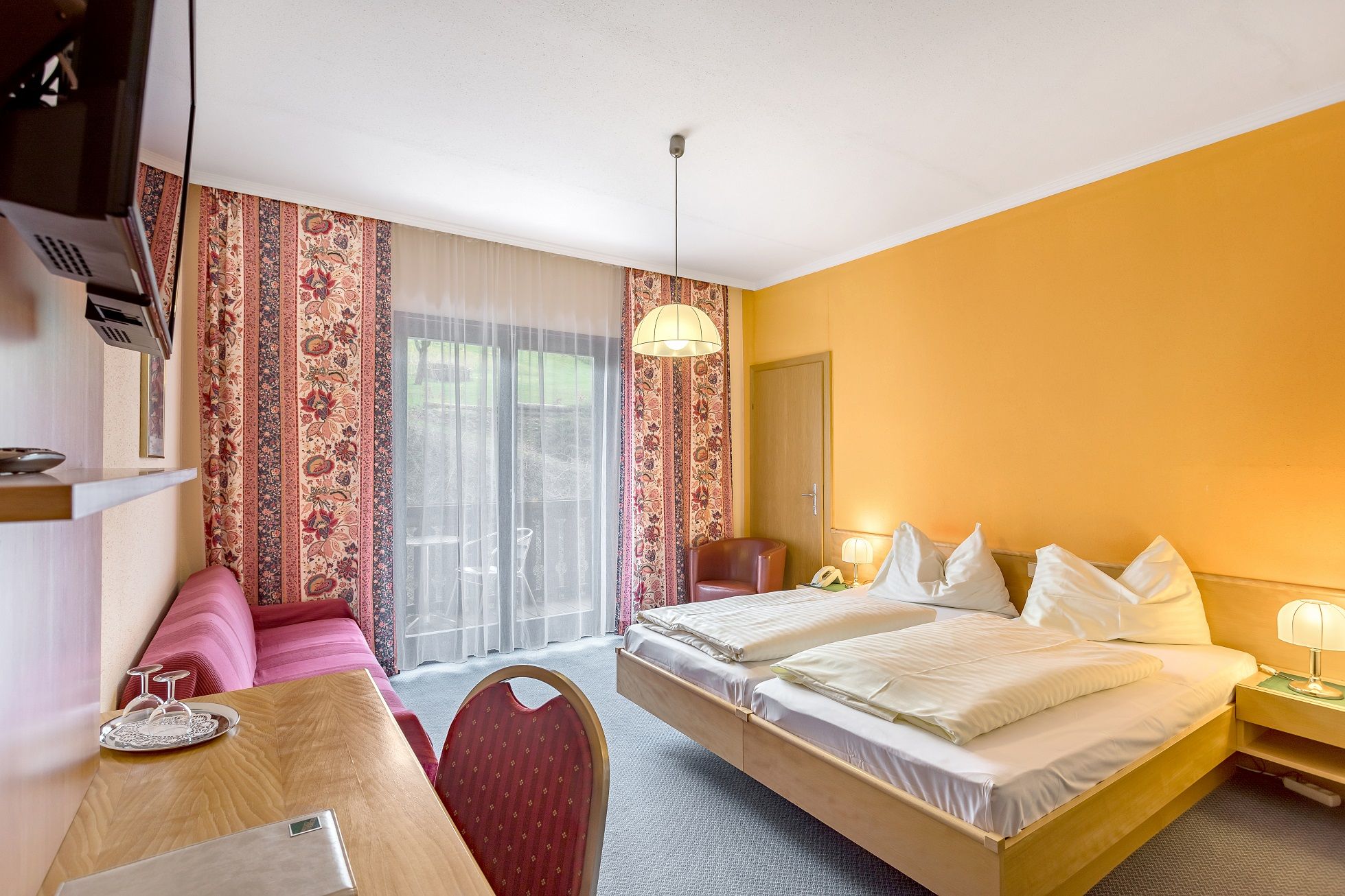 Hotelzimmer mit Doppelbett, gelben Wänden, rotem Sofa und Balkon.