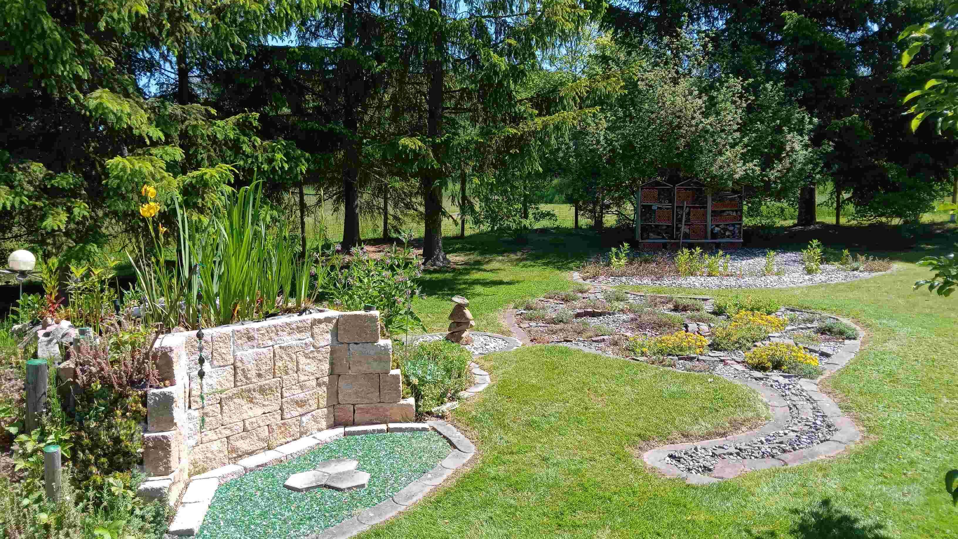 Ein gepflegter Garten mit Steinen, Pflanzen und einem Insektenhotel im Hintergrund.