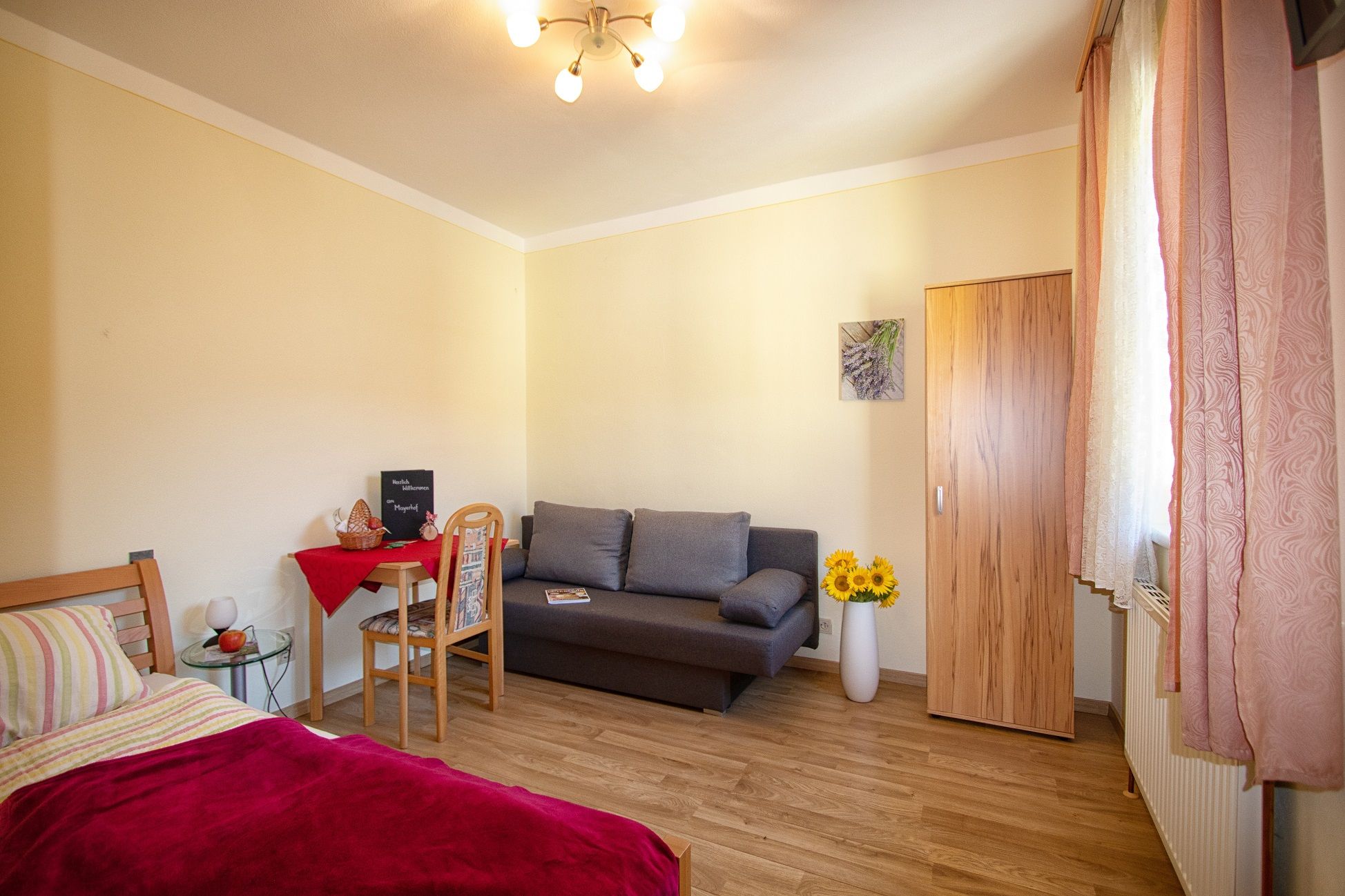 Gemütliches Zimmer mit Bett, Sofa, Tisch und Schrank, dekoriert mit Sonnenblumen und einem Bild an der Wand.