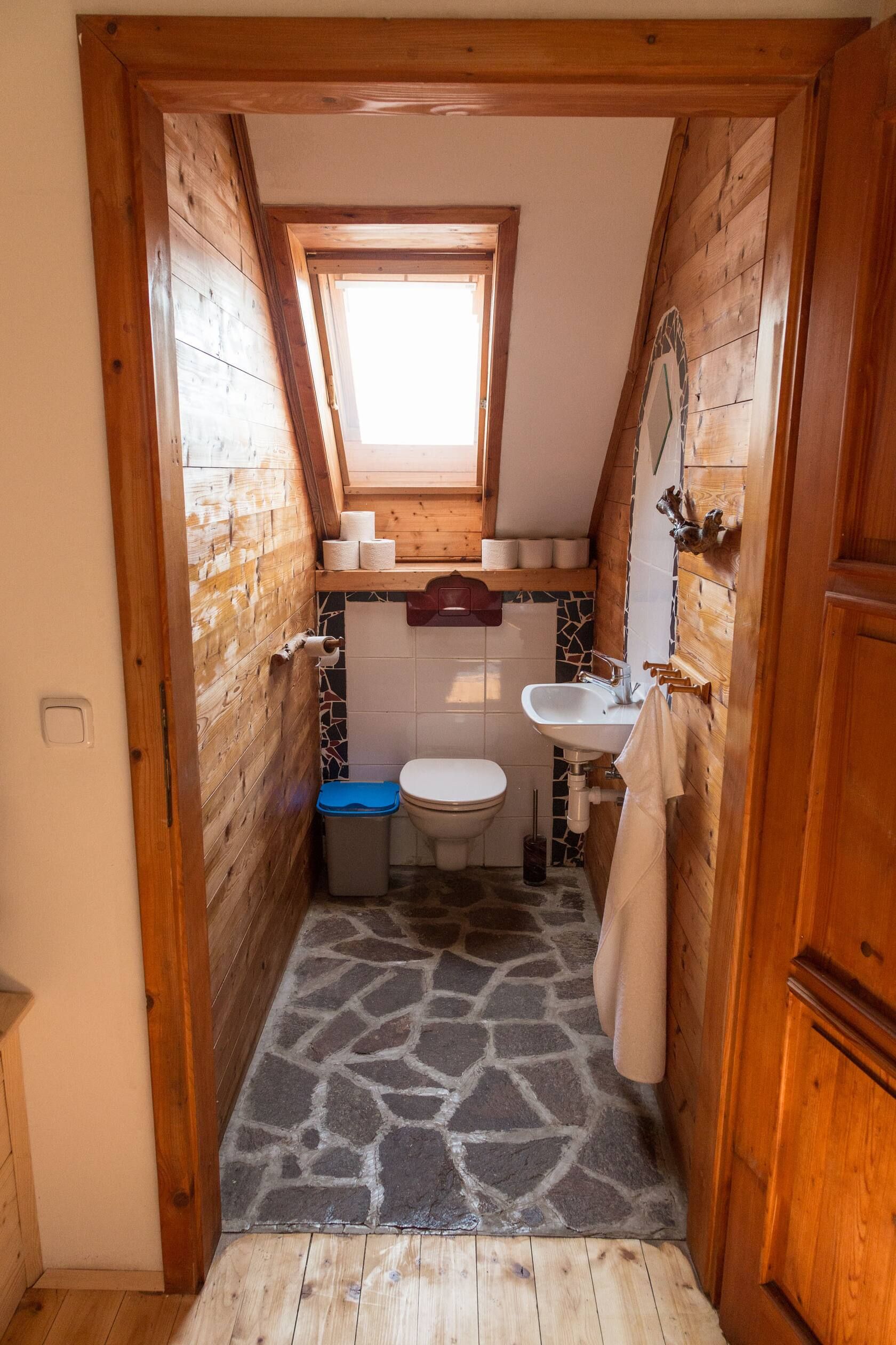 Kleines rustikales WC mit Holzverkleidung, Dachfenster, Toilette, Waschbecken und Steinboden.