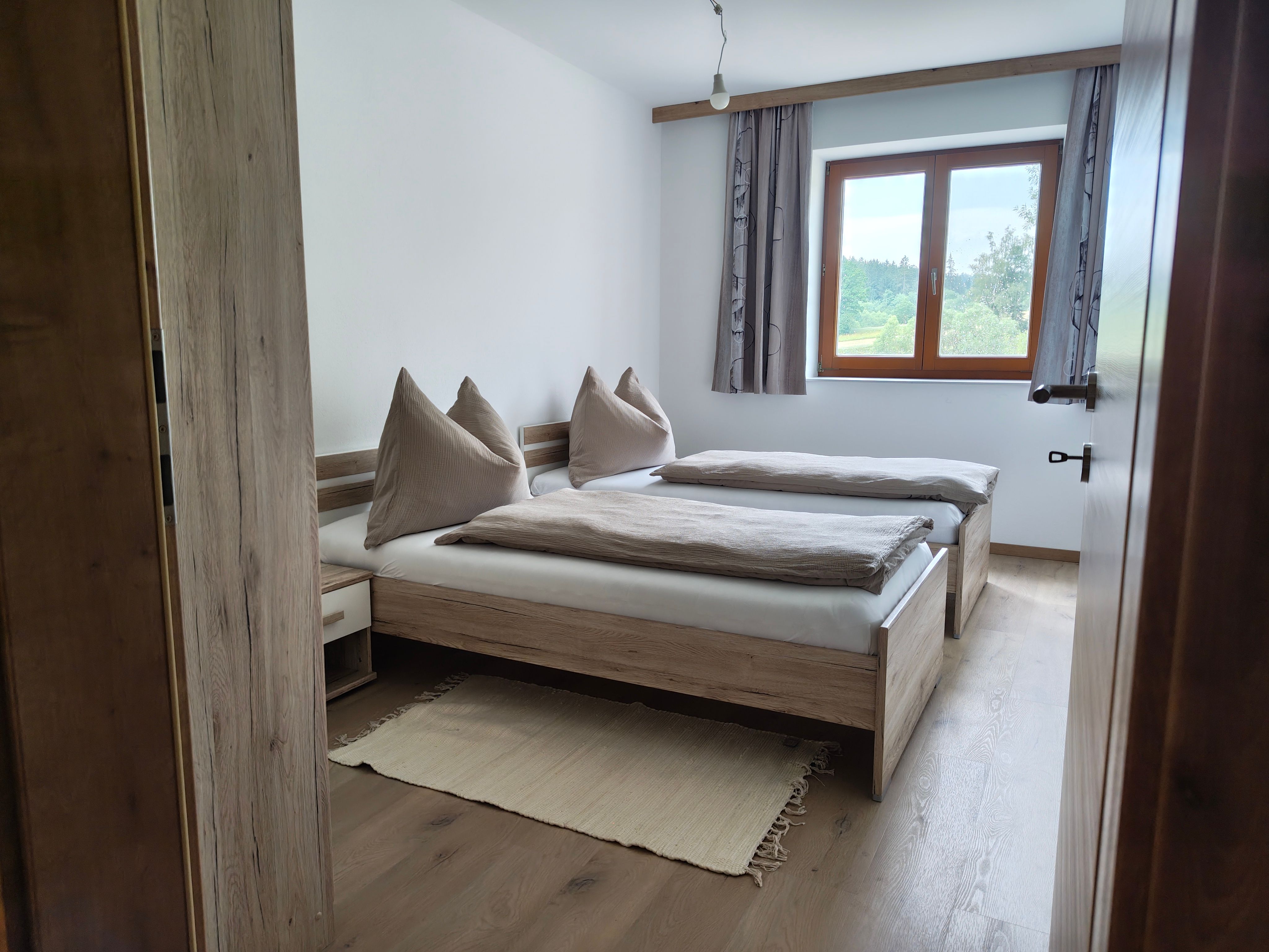 Helles Schlafzimmer mit Doppelbett aus Holz, beigen Kissen und Blick aus dem Fenster ins Grüne.