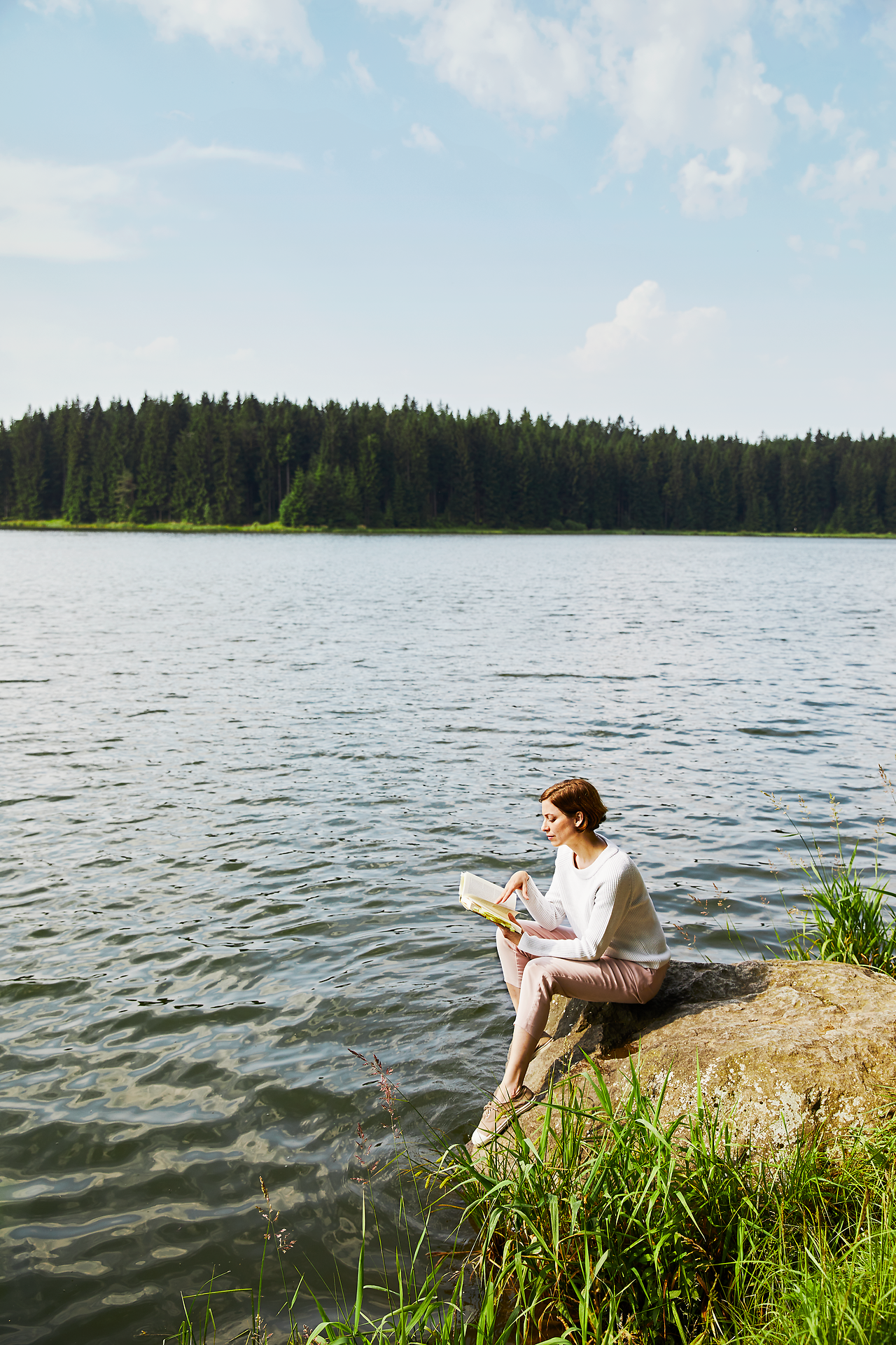 Am Ufer eines ruhigen Gewässers sitzt eine Frau entspannt auf einem großen Stein und vertieft sich in ein Buch. Umgeben von der idyllischen Natur des Waldviertels, strahlt die Szene eine friedliche Atmosphäre aus, die zum Verweilen und Träumen einlädt.