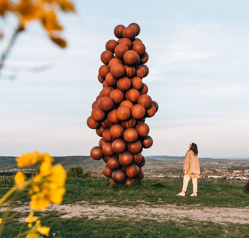 Eine beeindruckende Skulptur aus zahlreichen, rustikalen Kugeln erhebt sich majestätisch in die Höhe und spiegelt die kreative Verbindung zur Weinregion wider. Umgeben von sanften Hügeln und blühenden Wildblumen, lädt dieser Ort dazu ein, die Schönheit der Natur und die Kunst in harmonischem Einklang zu genießen.