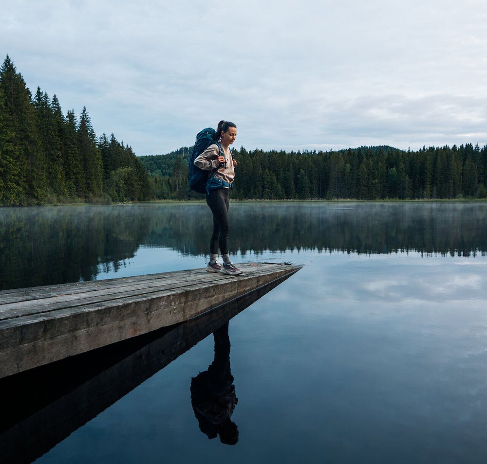 Frau steht auf einem Holzsteg am stillen Stierhübelteich, umgeben von dichter Waldlandschaft im Waldviertel, im Lichterspiel der Dämmerung.