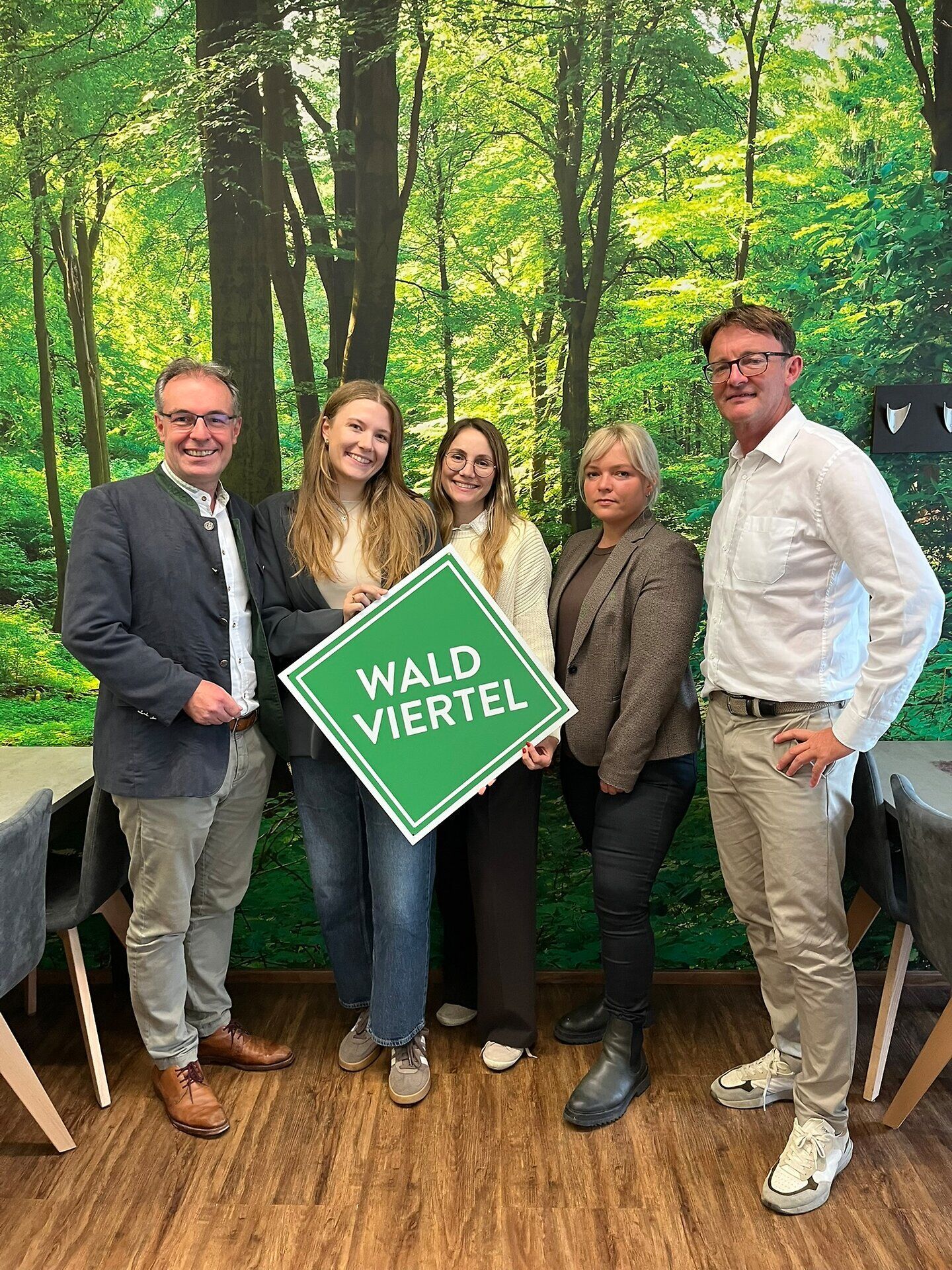 Fünf Personen stehen vor einer großflächigen Waldaufnahme. Zwei Personen halten gemeinsam ein grünes Schild mit der weißen Aufschrift „WALDVIERTEL“.