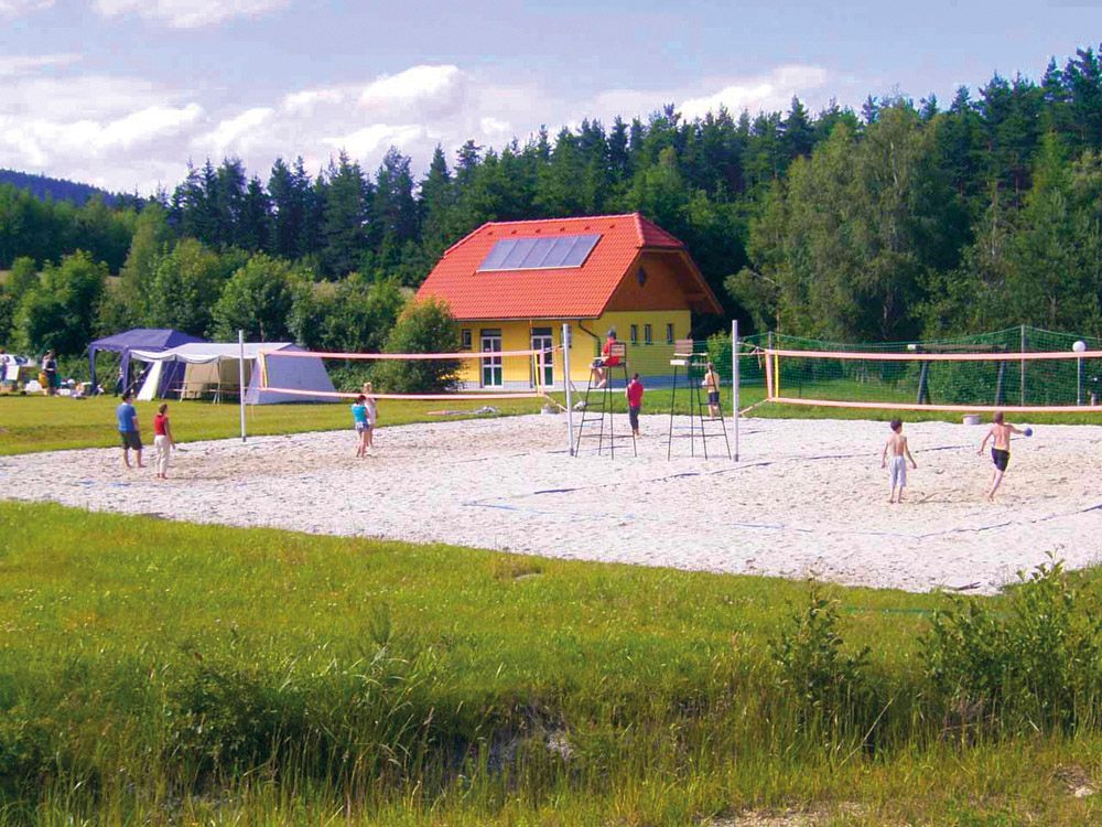 Menschen spielen Volleyball auf einem Sandplatz vor einem gelben Gebäude mit rotem Dach, umgeben von Bäumen.