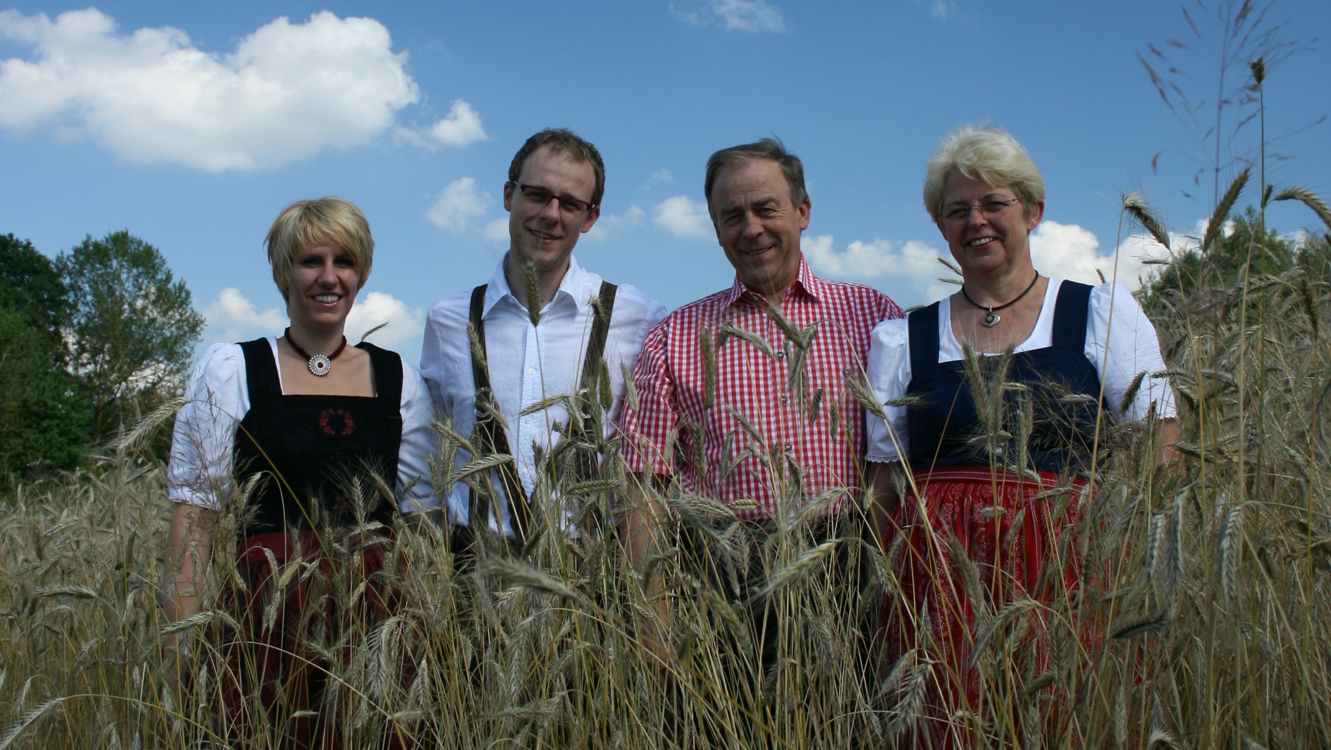 Vier Personen in traditioneller Kleidung stehen in einem Getreidefeld unter blauem Himmel.