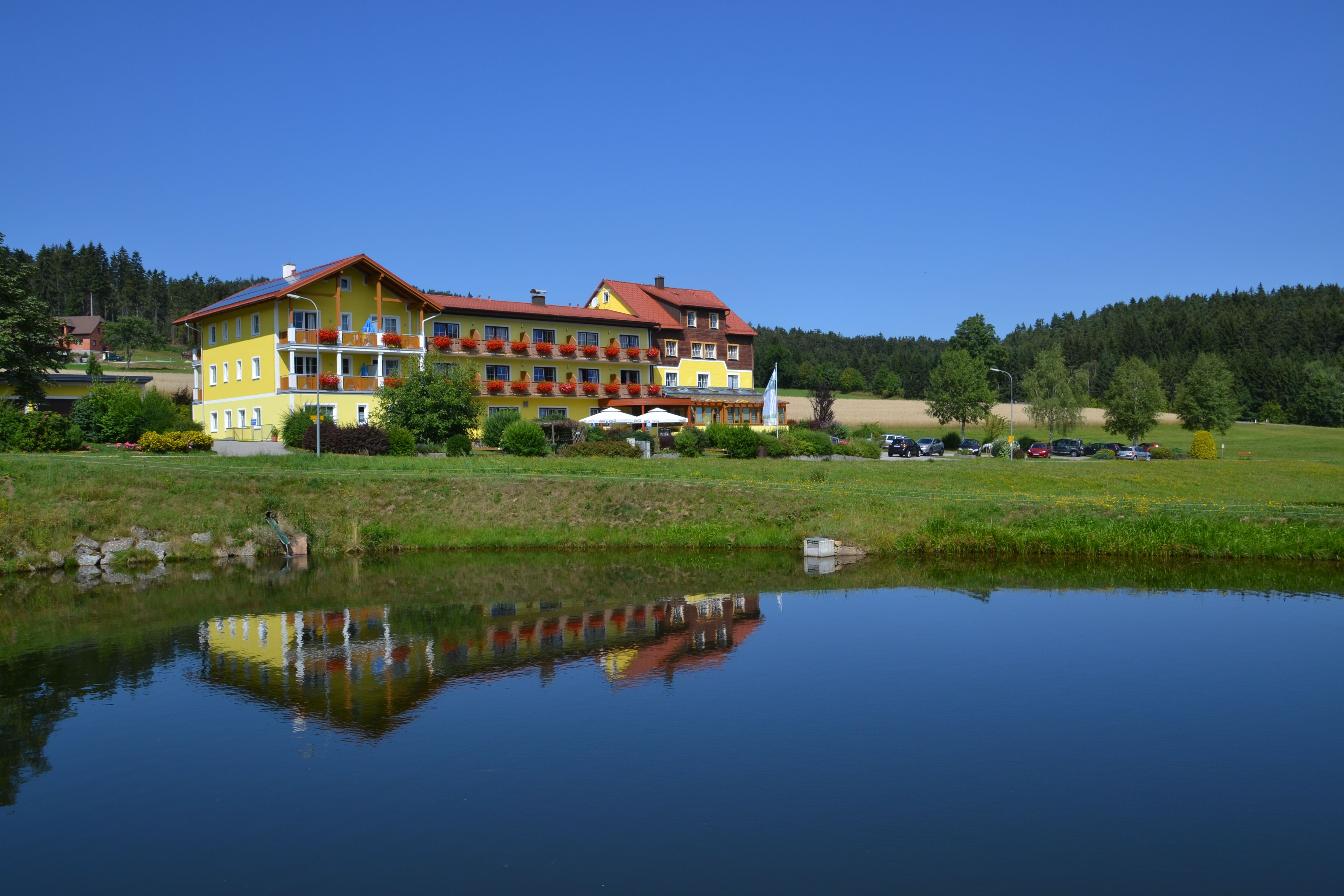 Ein gelbes Gebäude mit roten Dächern spiegelt sich in einem Teich, umgeben von grüner Landschaft und Bäumen.