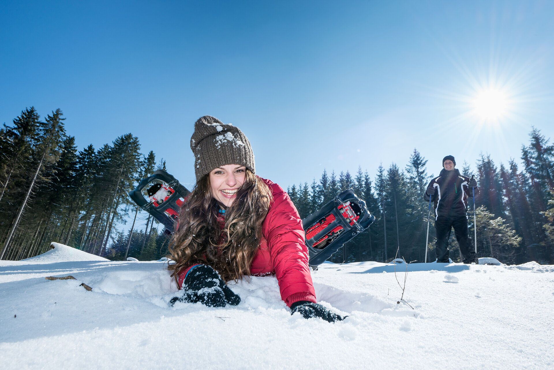 Zwei Personen im Schnee, eine liegt lachend im Vordergrund, die andere steht mit Schneeschuhen im Hintergrund. Sonniger Wintertag mit Wald im Hintergrund.