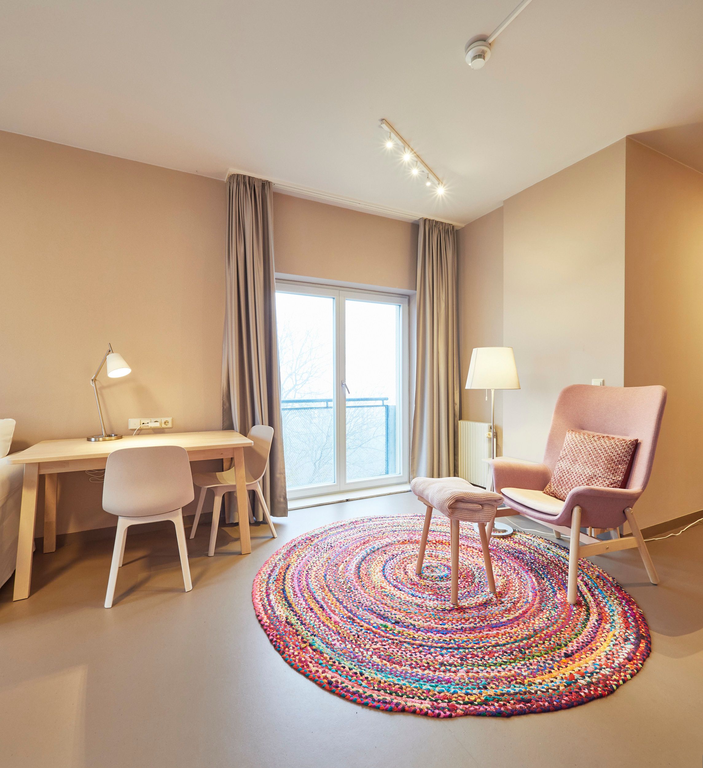 Helles Zimmer mit Schreibtisch, Stuhl, Sessel und buntem Teppich.