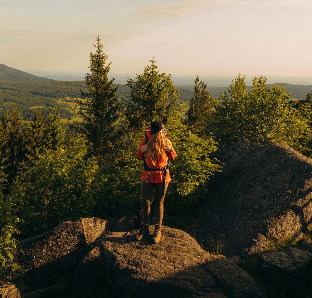 Ein Wanderer steht auf einem Felsen mit Ausblick auf bewaldete Hügel, aufgenommen im Waldviertel, gekrönt von einem klaren Himmel und dem Licht der aufgehenden Sonne.