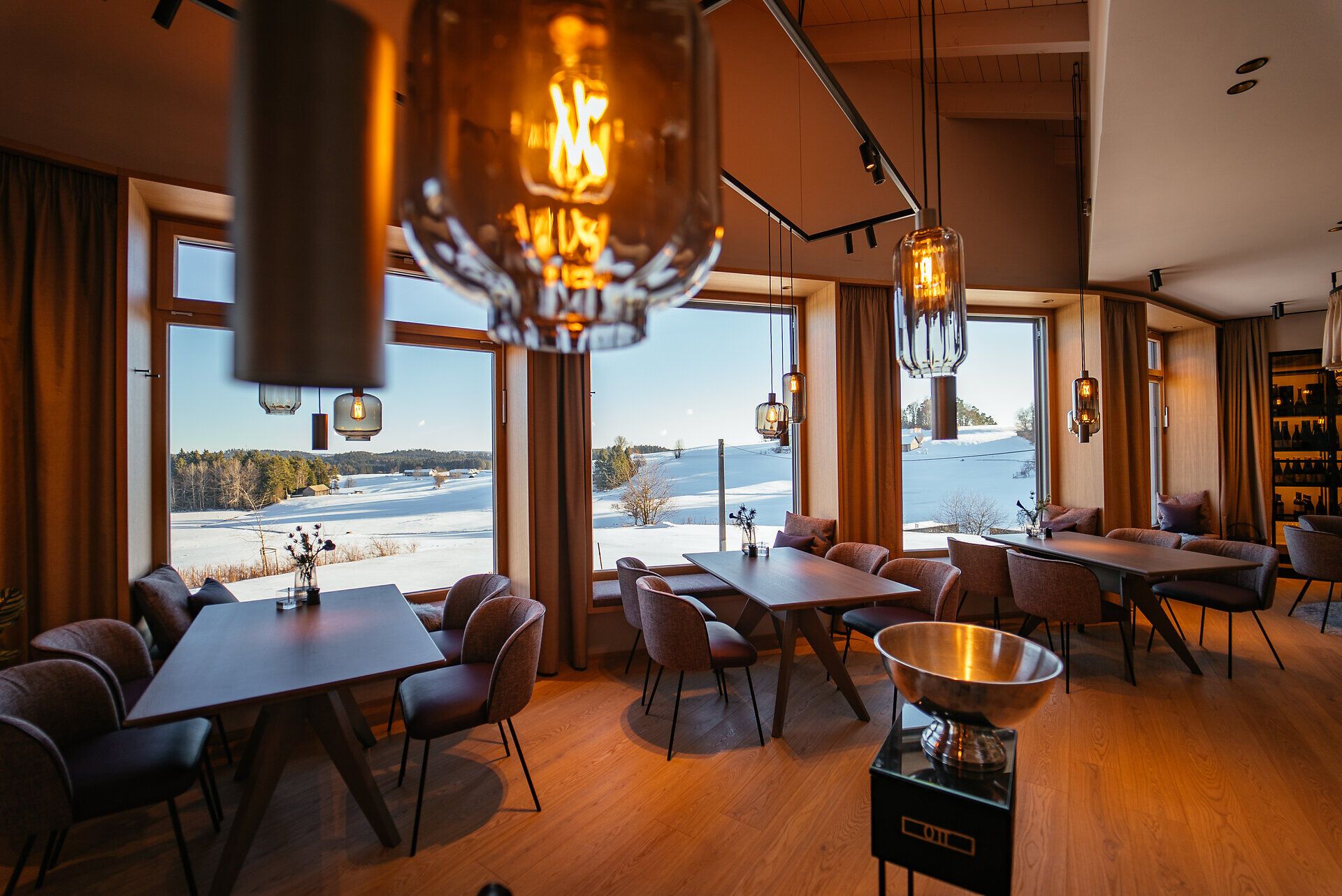 In einem stilvoll eingerichteten Restaurant mit großen Fenstern, die einen atemberaubenden Blick auf die verschneite Landschaft bieten, lädt eine gemütliche Atmosphäre zum Verweilen ein. Die warmen Lichtakzente und die geschmackvolle Einrichtung schaffen einen einladenden Raum, ideal für genussvolle Stunden mit regionalen Köstlichkeiten.