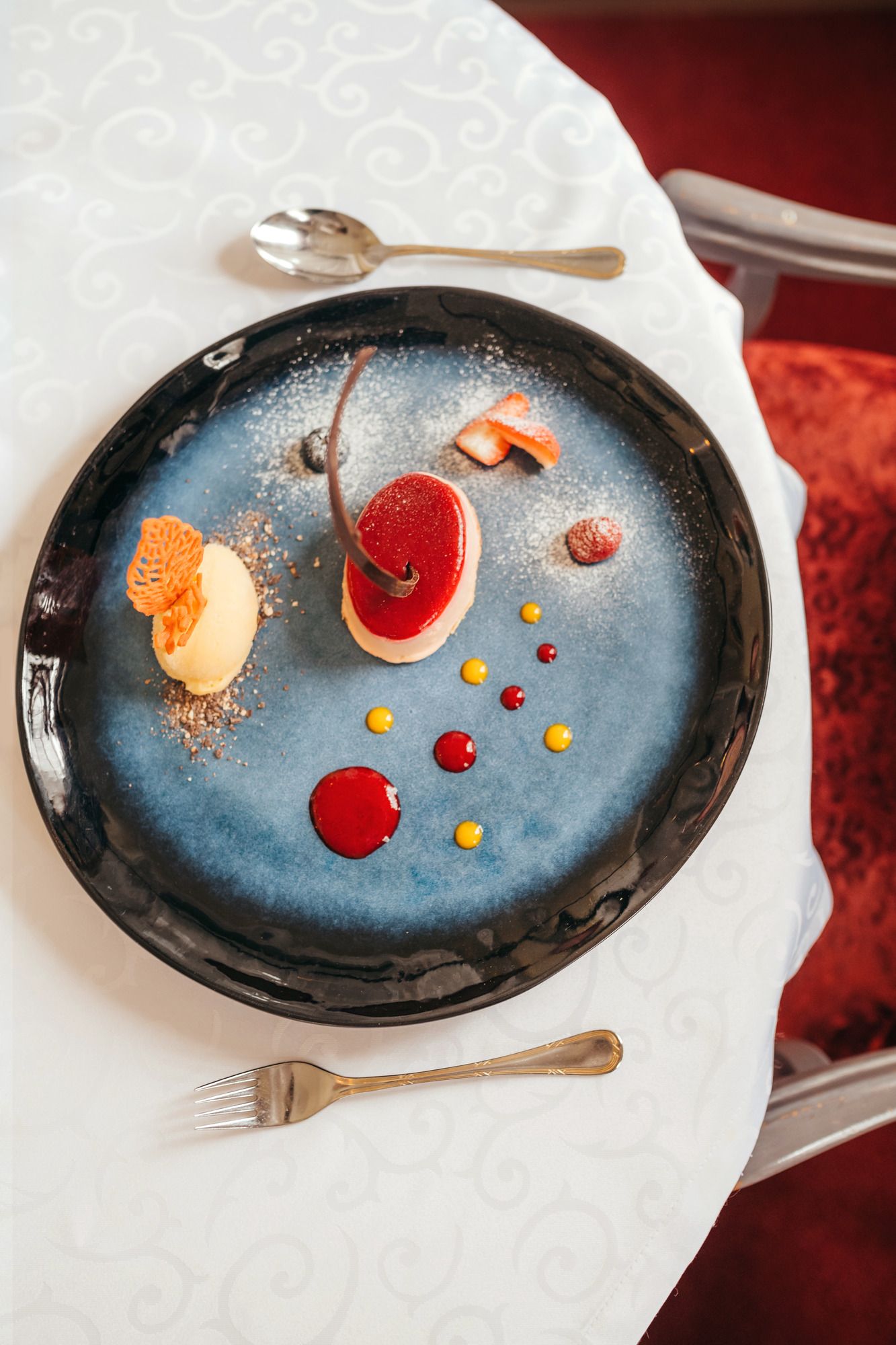 Ein kunstvoll angerichtetes Dessert auf einem blauen Teller mit roten und gelben Saucenpunkten.