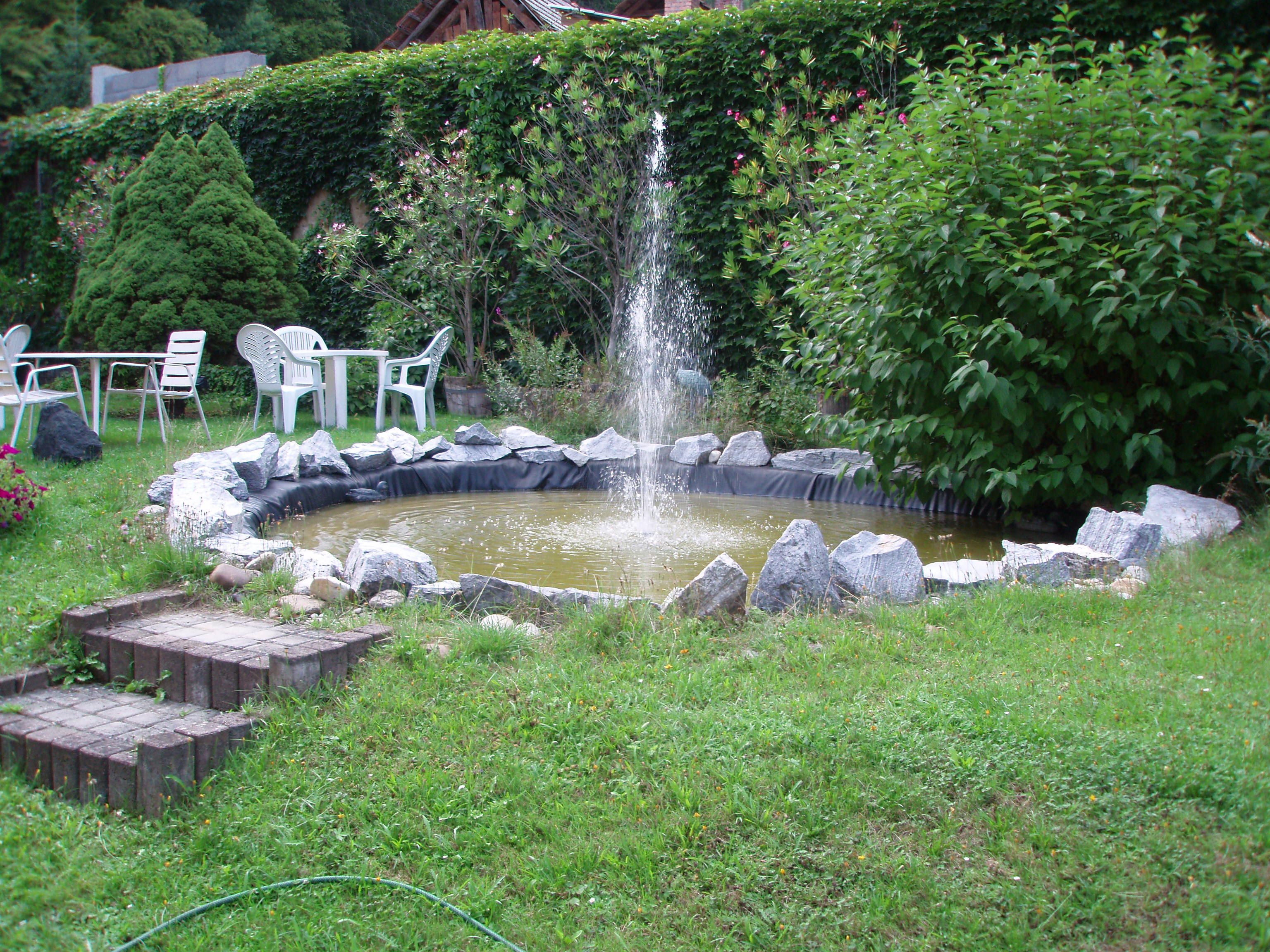 Garten mit kleinem Teich und Springbrunnen, umgeben von Pflanzen und Gartenmöbeln.