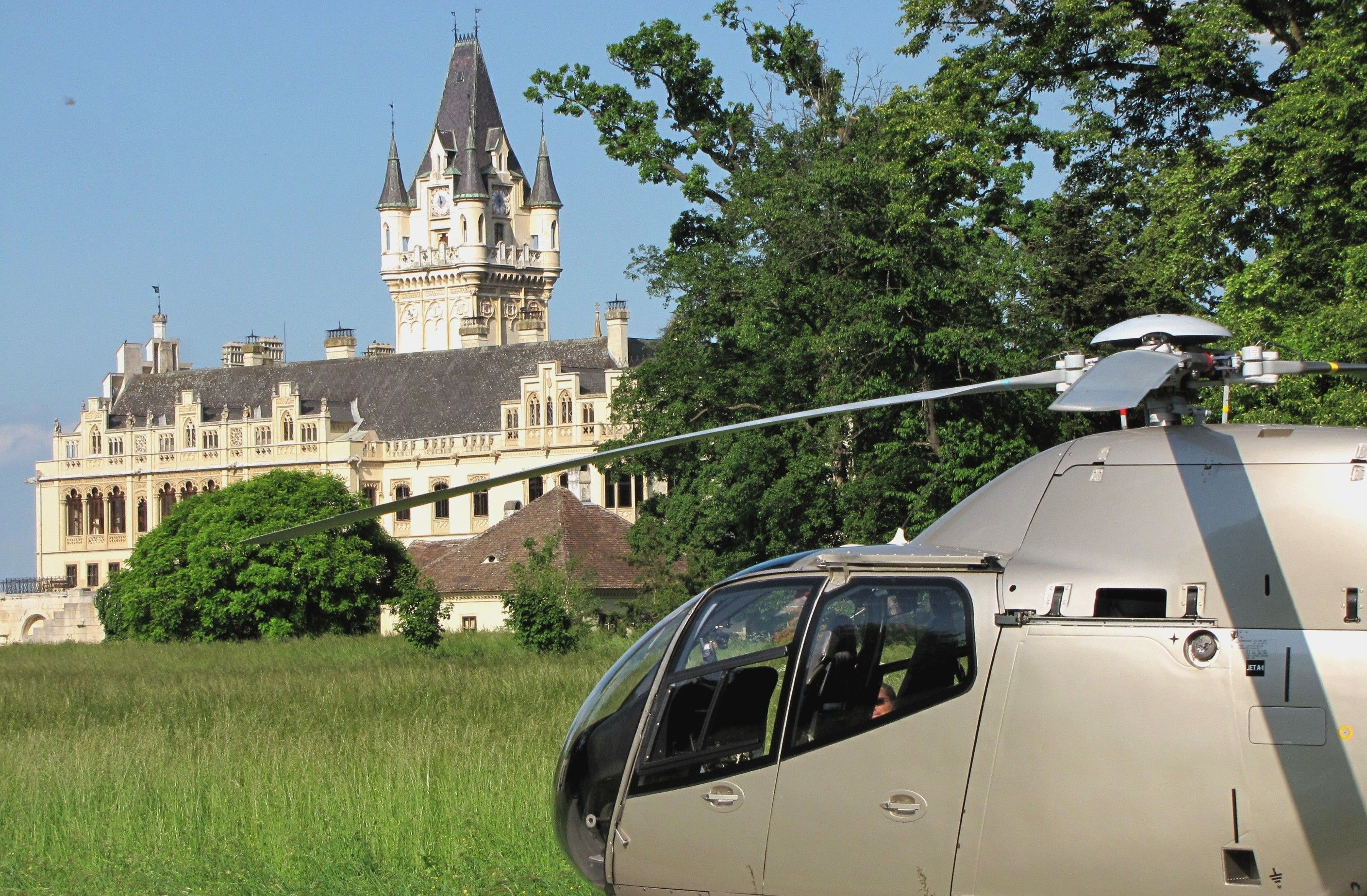 Ein Helikopter steht auf einer Wiese vor einem großen, historischen Schloss mit Türmen und einem Uhrturm.