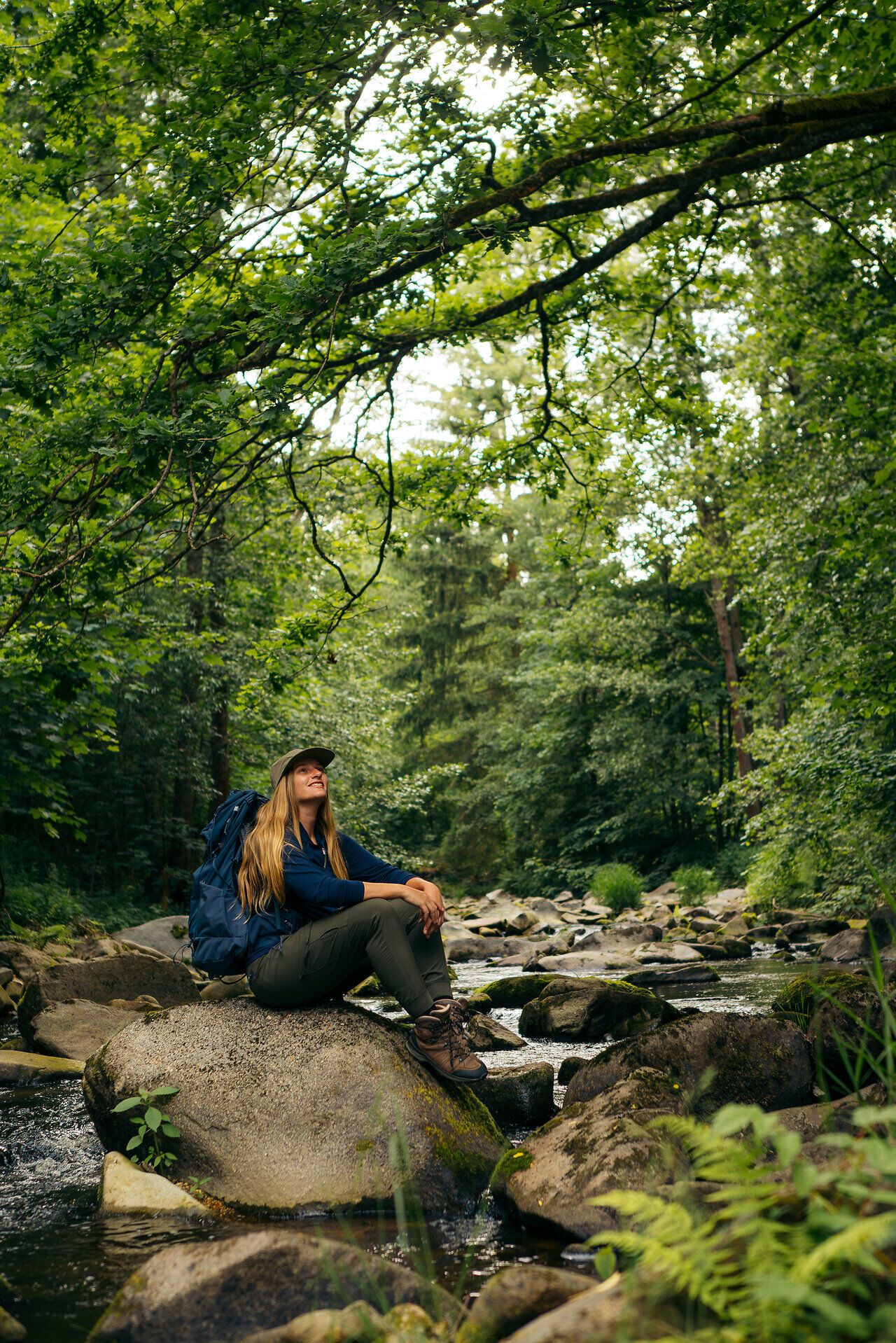 Eine Frau mit Rucksack sitzt auf einem Felsen in einem Bach im Gabrielental, umgeben von üppigem Grün, im Waldviertel.
