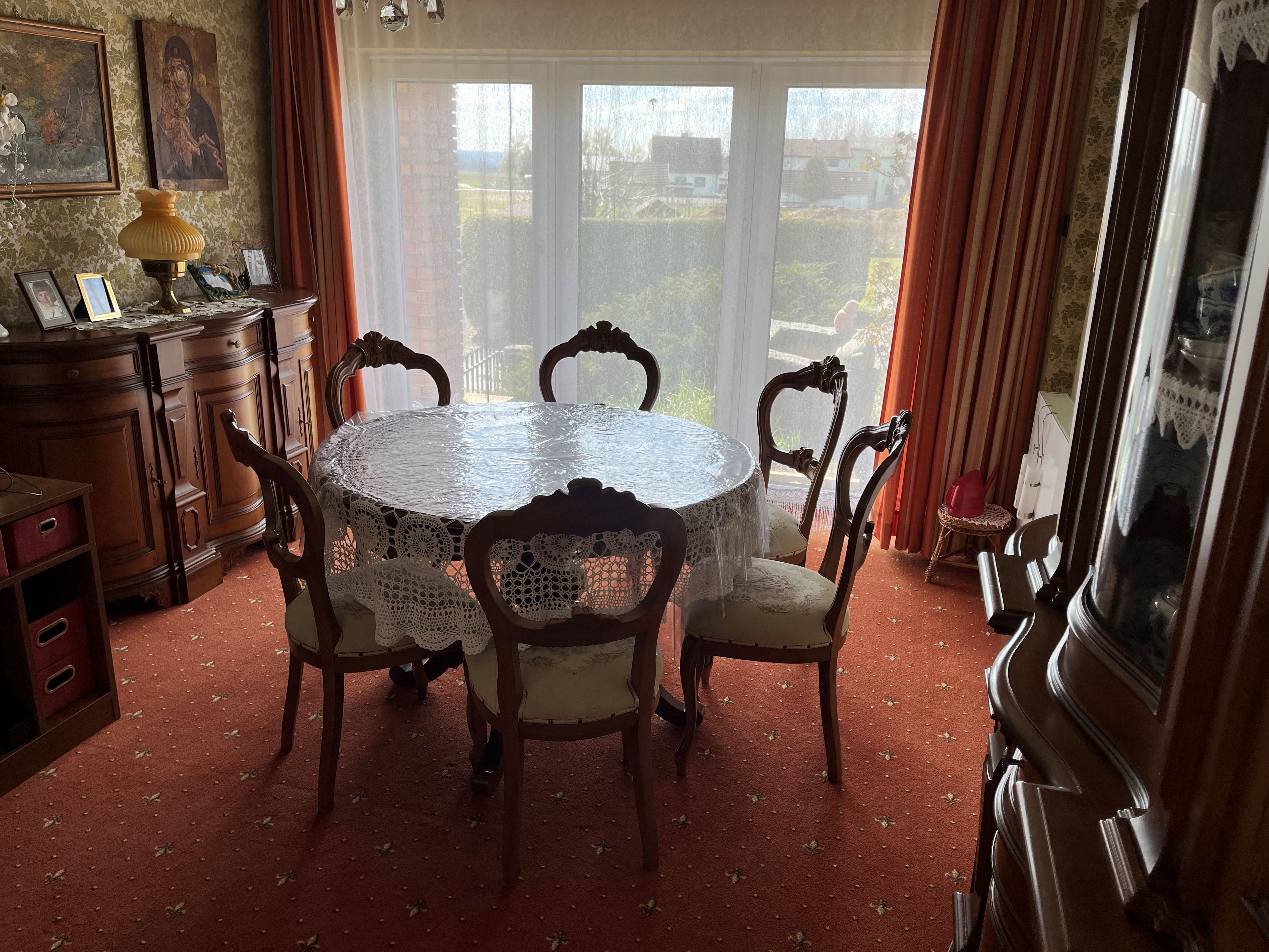 Ein traditionell eingerichtetes Esszimmer mit einem runden Tisch, der mit einer Spitzen-Tischdecke bedeckt ist, und sechs Stühlen. Im Hintergrund sind große Fenster mit Vorhängen zu sehen.