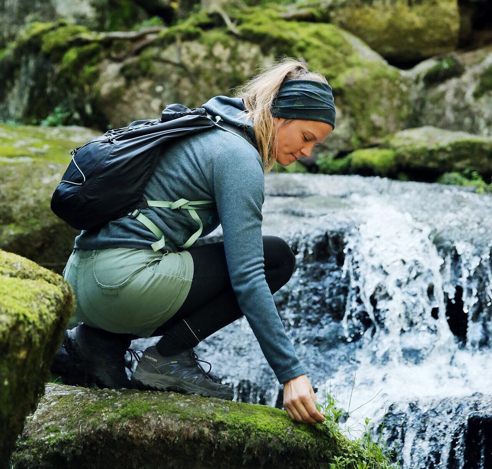 Inmitten der üppigen Natur kniet eine Wanderin am Ufer eines plätschernden Baches und bewundert die frische, grüne Umgebung. Die sanften Wasserfälle im Hintergrund verleihen der Szenerie eine magische Atmosphäre, die zum Verweilen einlädt. Hier, wo die Ruhe der Natur auf die Schönheit des Wassers trifft, wird jeder Moment zum unvergesslichen Erlebnis.