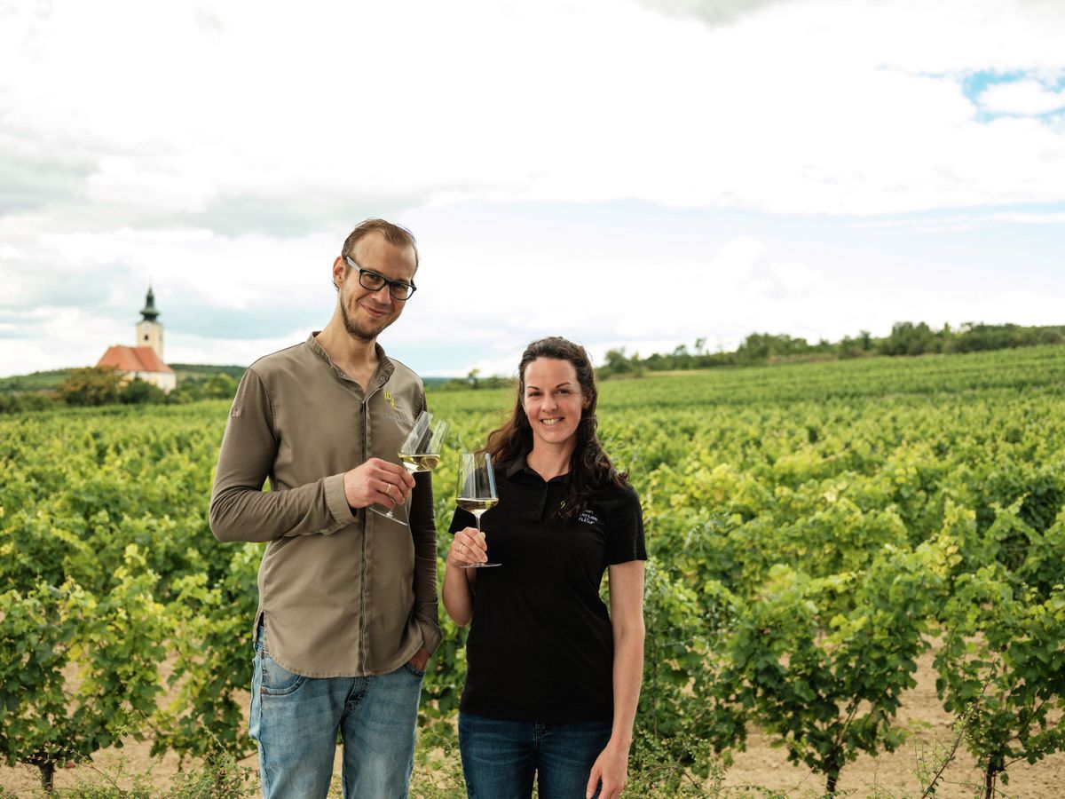 Zwei Personen stehen mit Weingläsern in einem Weinberg, im Hintergrund eine Kirche.