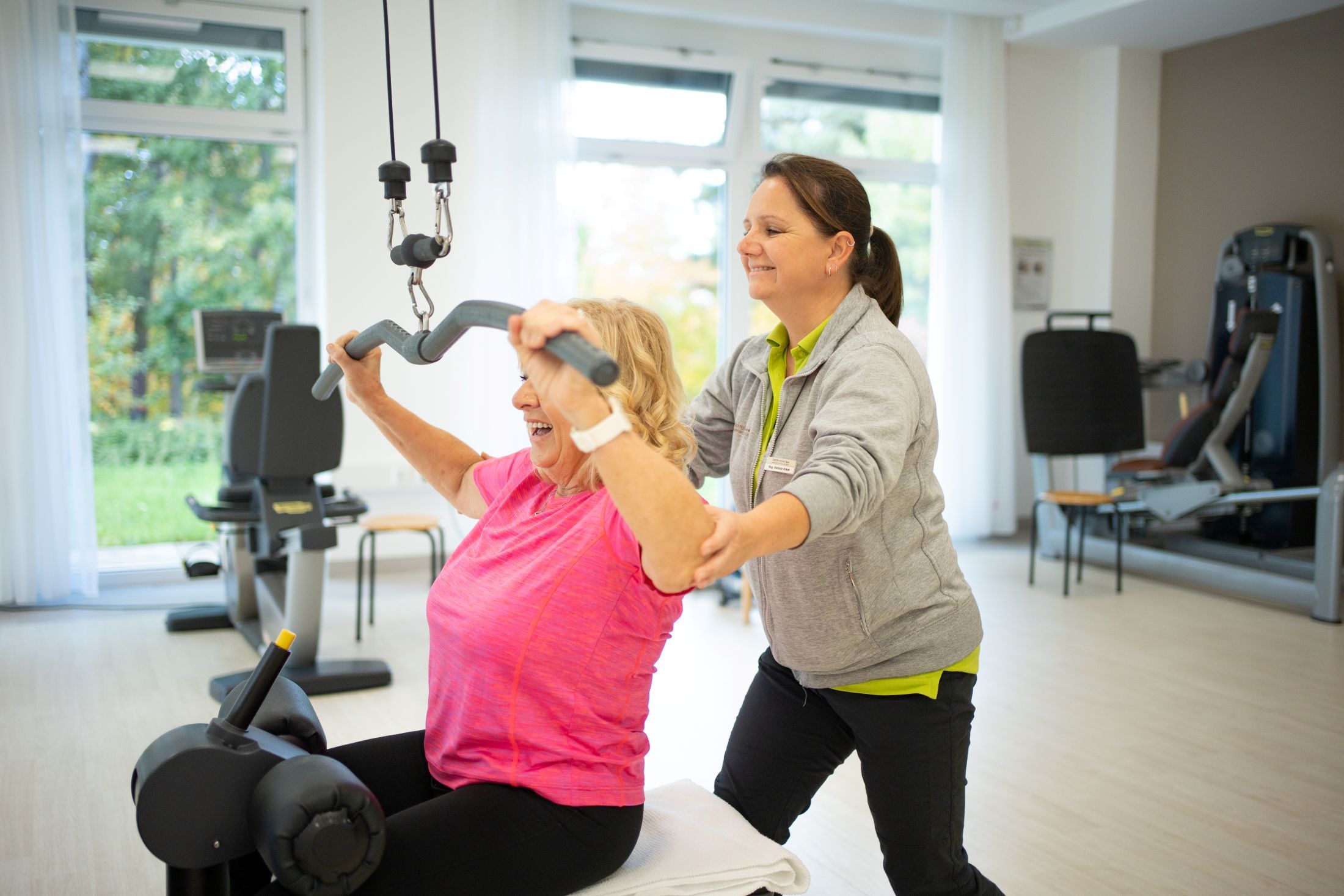 Eine ältere Frau trainiert an einem Fitnessgerät, unterstützt von einer Trainerin in einem hellen Raum.