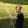 Mann mit Angelrute am Teich vor grüner Landschaft.
