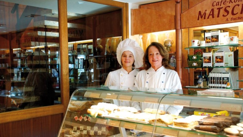 Zwei Frauen in Konditoruniformen stehen hinter einer Theke mit Gebäck in einer Bäckerei.