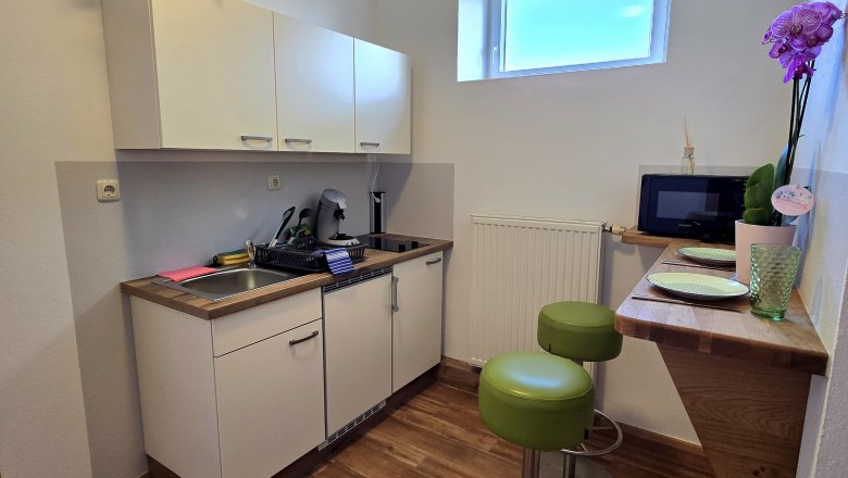 Small kitchen with white cupboard, sink, two green bar stools and small window.
