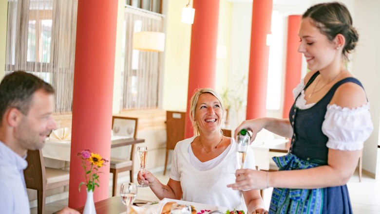 Eine Frau in Tracht schenkt einem Paar in einem Restaurant Sekt ein.