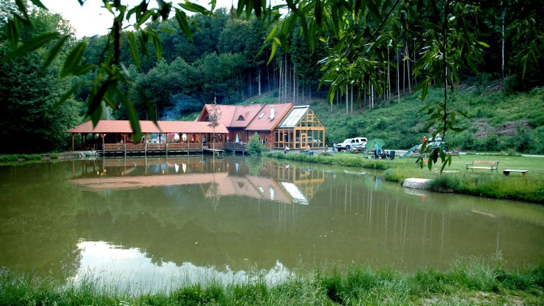 A building with a red roof is reflected in a pond surrounded by trees and meadows.