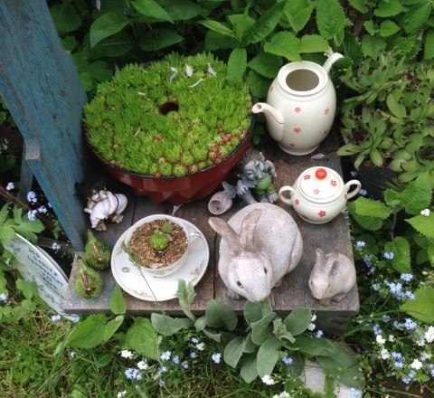 Gartenarrangement mit Teekanne, Tasse, Hasenfiguren und Sukkulenten.