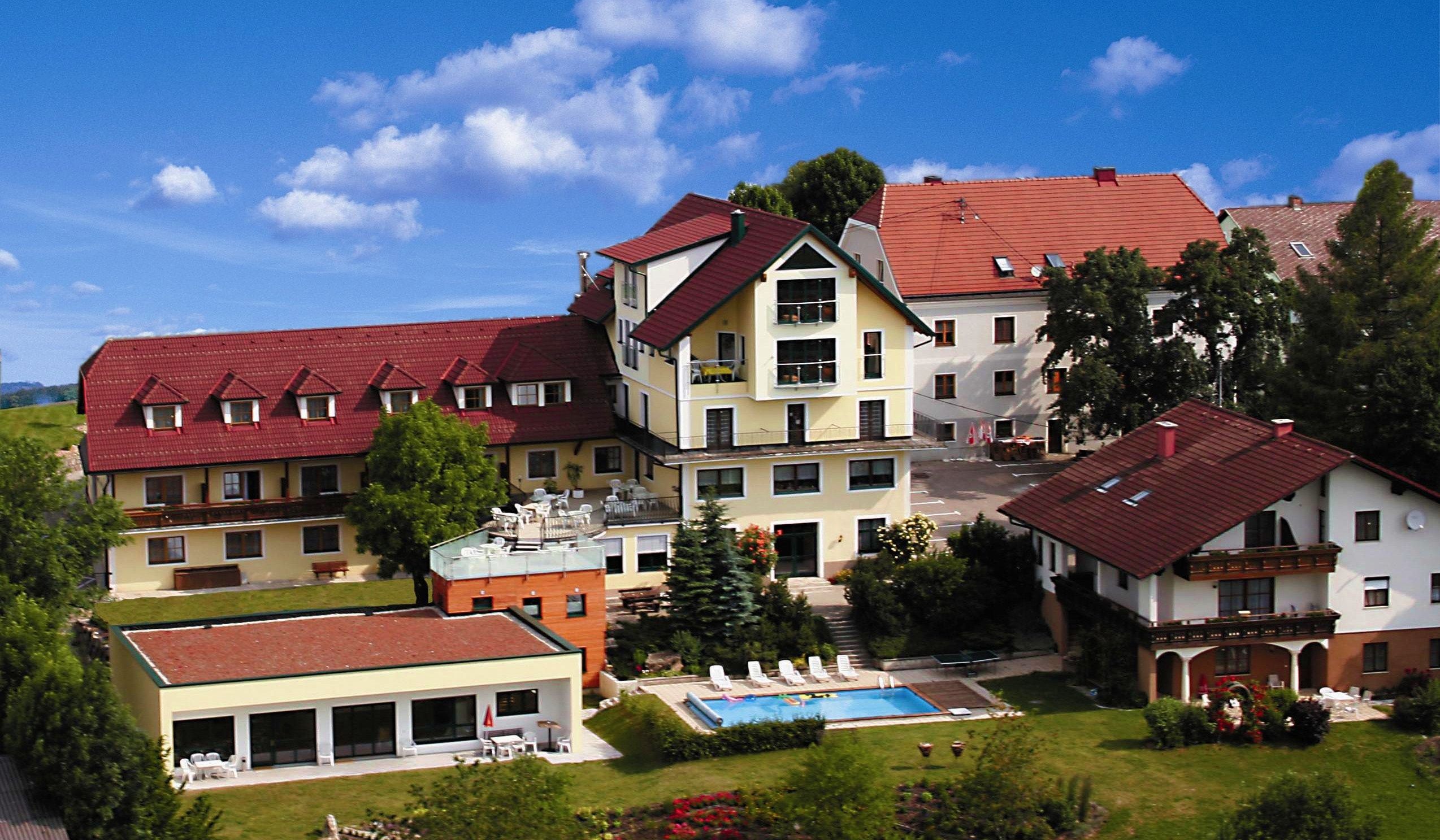 Ein Hotelkomplex mit roten Dächern, Pool und Garten, umgeben von Bäumen und blühenden Pflanzen unter einem blauen Himmel.