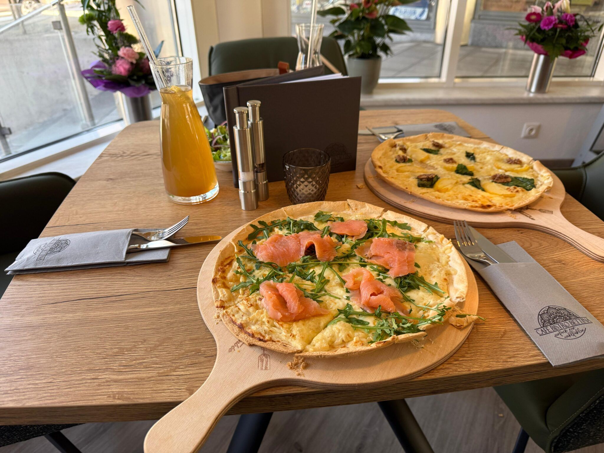 Zwei Flammkuchen auf Holzbrettern auf einem Tisch in einem Café, mit Getränkekaraffe und Blumen im Hintergrund.