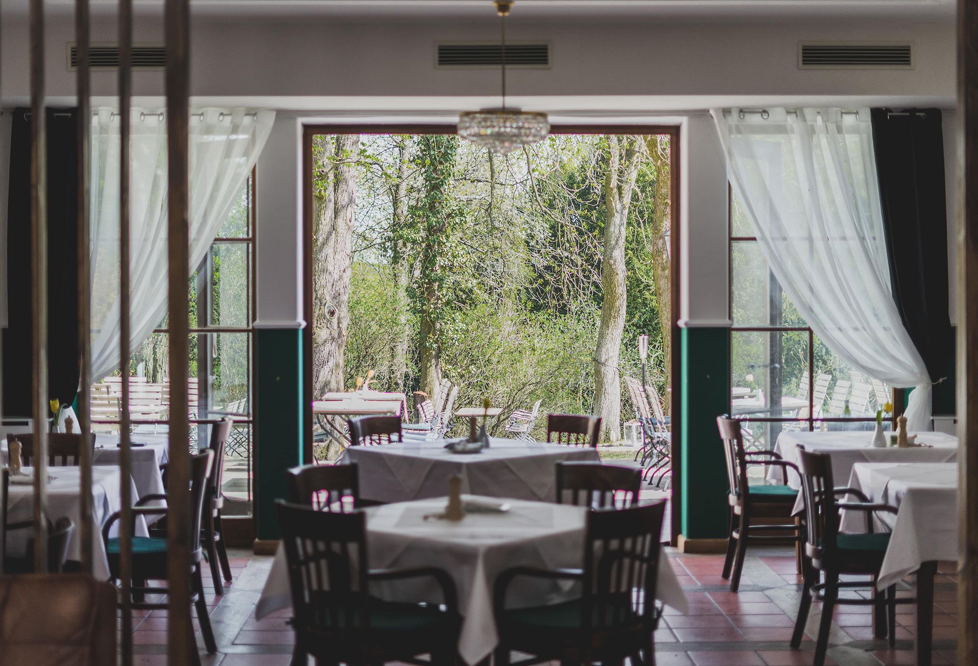 Ein Restaurant mit gedeckten Tischen und Blick auf einen grünen Garten durch große Fenster.