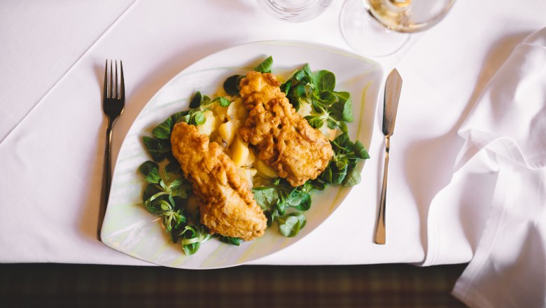 Ein Teller mit gebackenem Karpfenfilet auf Feldsalat und Kartoffeln, auf einem weiß gedeckten Tisch.