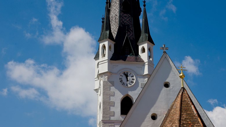 Kirchturm mit Uhr und spitzem Dach vor blauem Himmel.