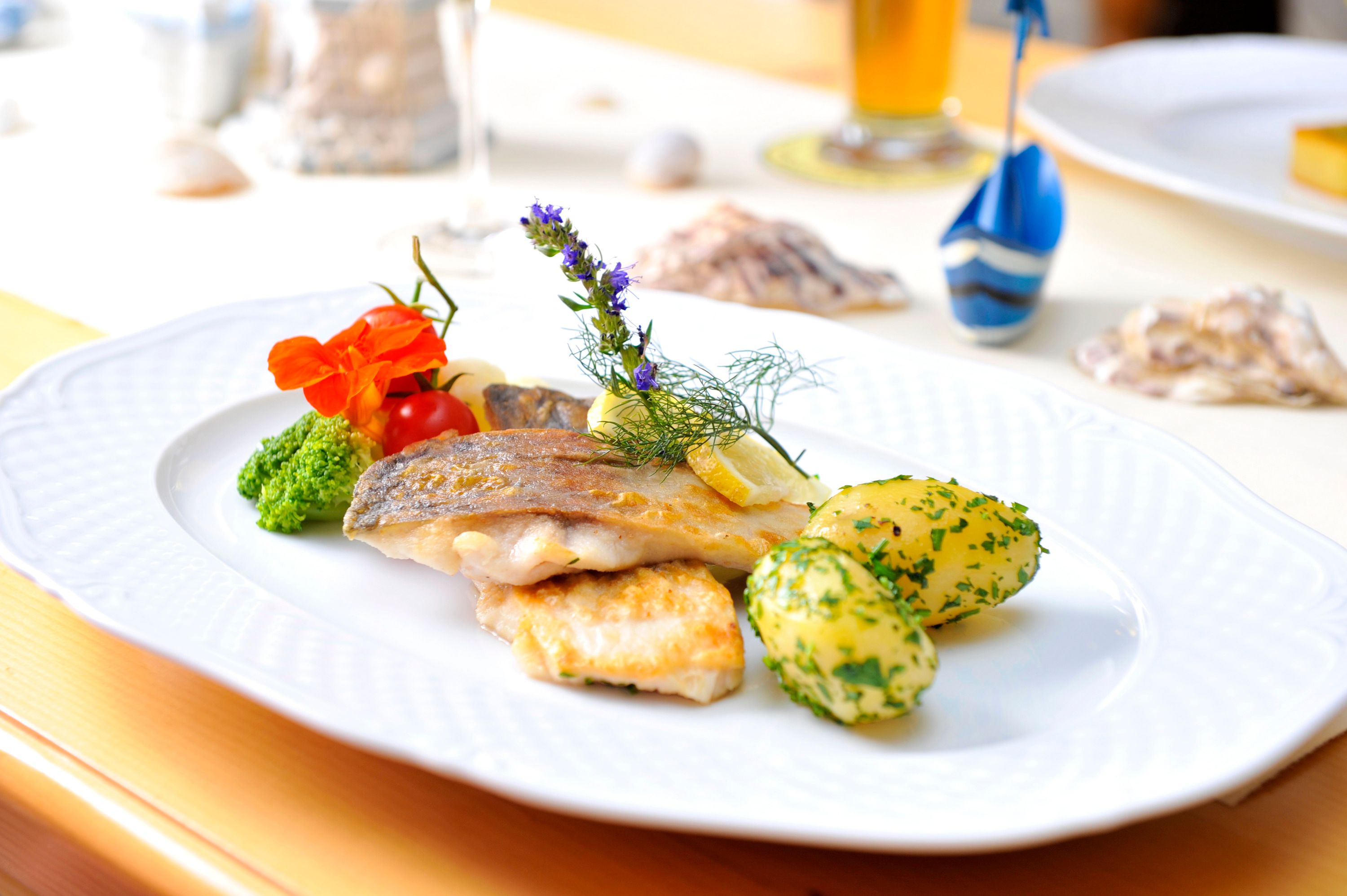Ein Teller mit gebratenem Fischfilet, Kartoffeln und Gemüse, dekoriert mit Kräutern und Blüten.