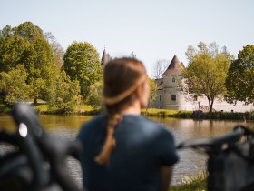 Ein sanfter Wind weht &uuml;ber das glitzernde Wasser, w&auml;hrend die B&auml;ume in sattem Gr&uuml;n erstrahlen. Die malerische Kulisse des Schlosses l&auml;dt Radfahrer ein, eine wohlverdiente Pause einzulegen und die Sch&ouml;nheit der Natur zu genie&szlig;en.
