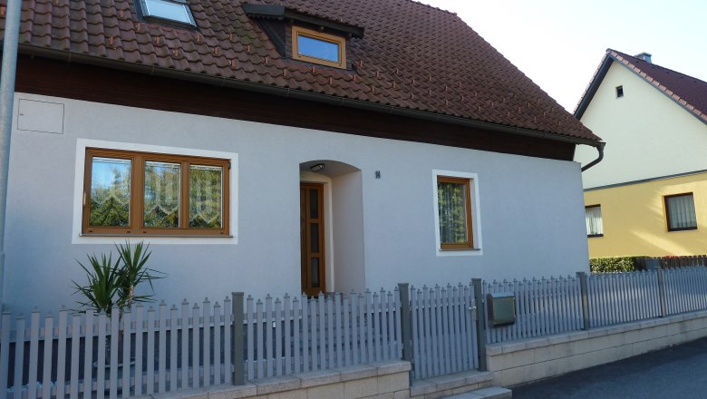 Ein Einfamilienhaus mit grauer Fassade, braunem Dach und Holztüren. Ein kleiner Zaun umgibt das Grundstück.