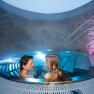 A couple relaxes in a round whirlpool with blue lighting and stone walls.